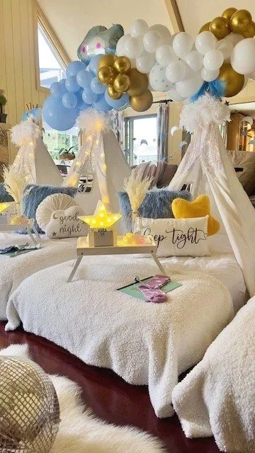 Decorated room with balloon arch in blue, white, gold, and silver, white teepees with fluffy accents, plush pillows including a star-shaped one, a small star-shaped lamp, and a blanket with 'sleep tight' embroidered, creating a cozy, festive setting for a celebration or gathering.