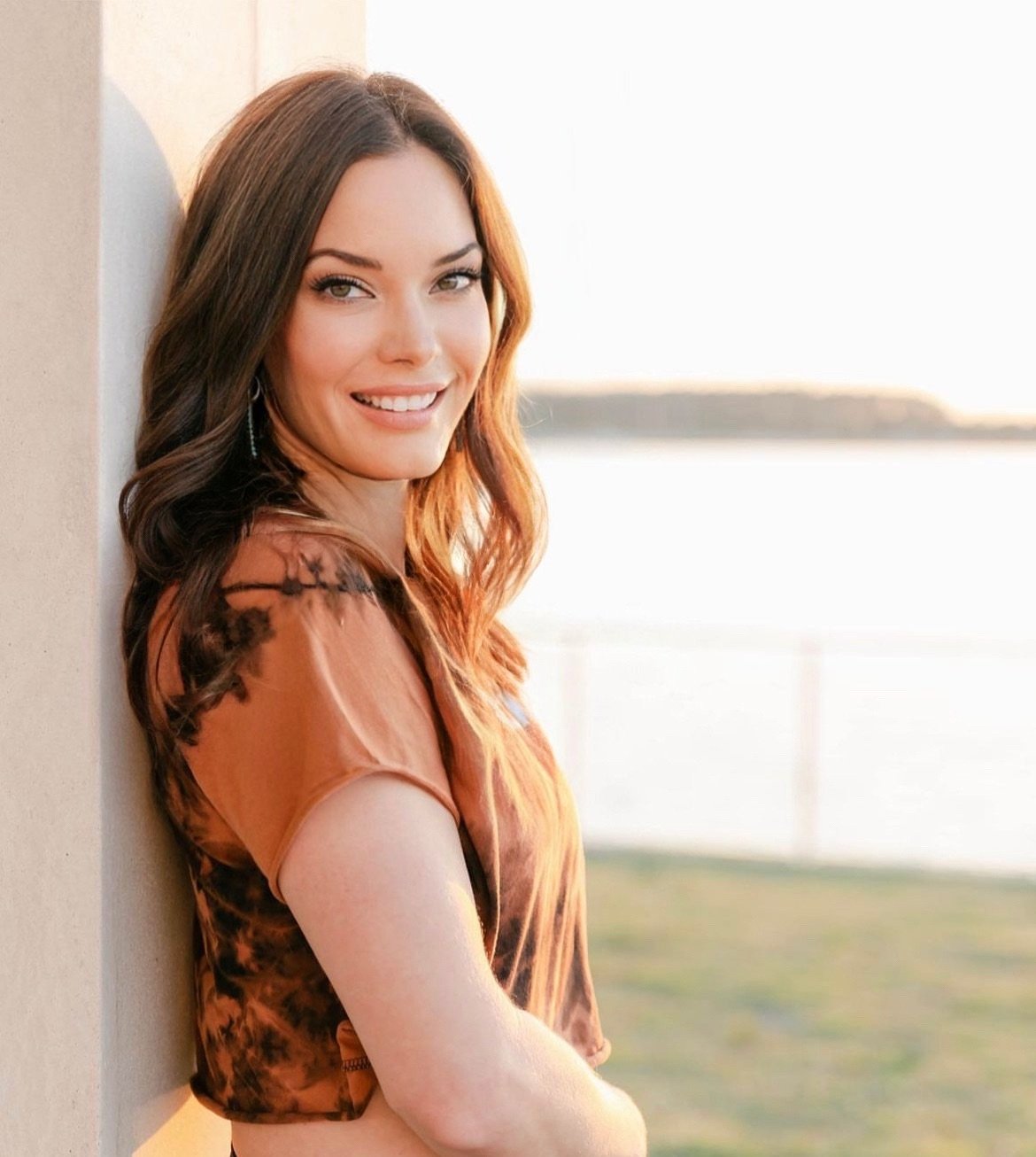 Smiling woman with long hair leaning against a wall, wearing a tie-dye top and earrings, outdoors at sunset.