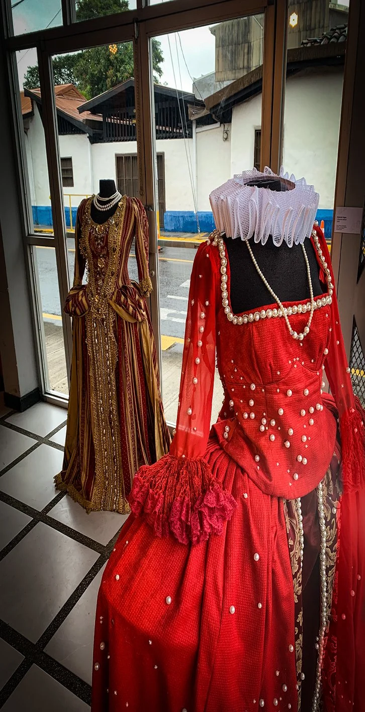 Dos vestidos de fiesta con accesorios, en una vitrina, uno rojo con perlas y encajes, y uno con rayas doradas y rojas, en un entorno interior junto a ventanas.
