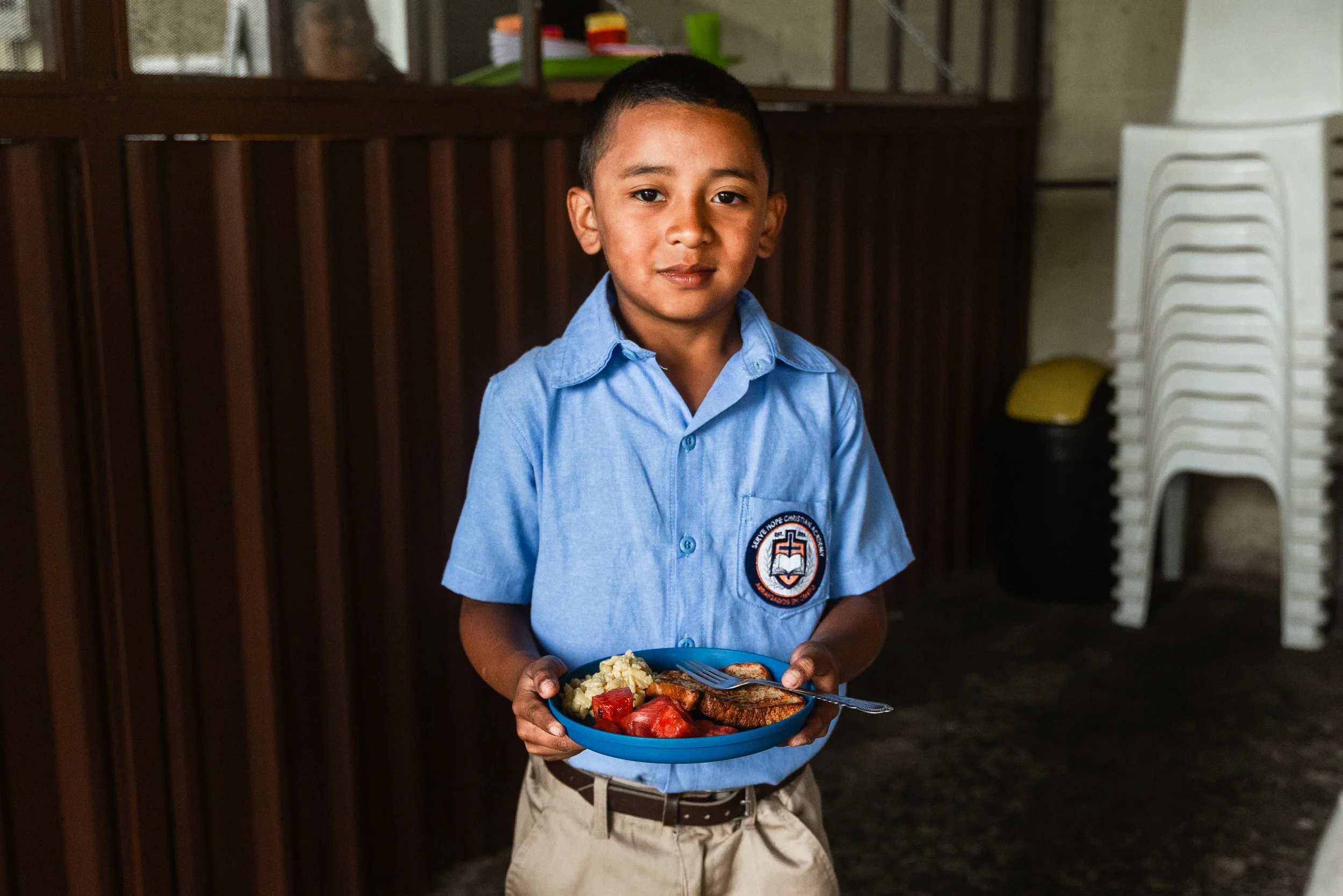 Young boy in a blue uniform smiles while holding a plate of nutritious food at Serve Hope Christian Academy, highlighting the school's commitment to combating food insecurity through daily breakfast and lunch.