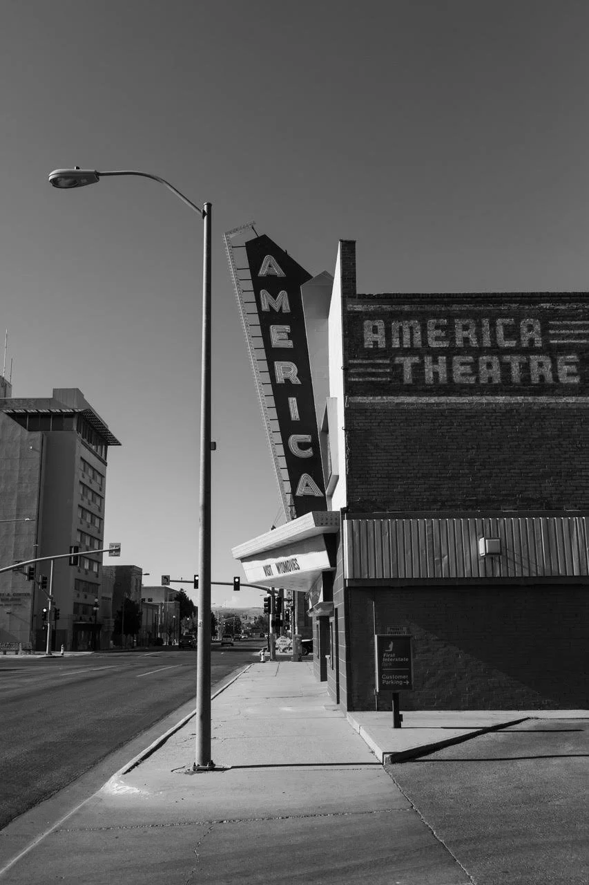 Street view of the American Theatre with a vertical marquee sign and a brick building with the words 'AMERICA THEATRE' on it, in black and white by Amy Sacka.