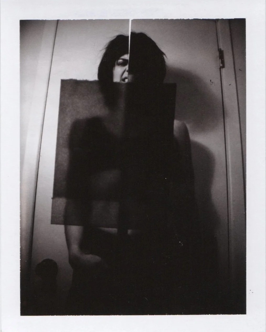 Black and white photo of a person taking a mirror selfie in a room with a door in the background, with a partial reflection of their face visible