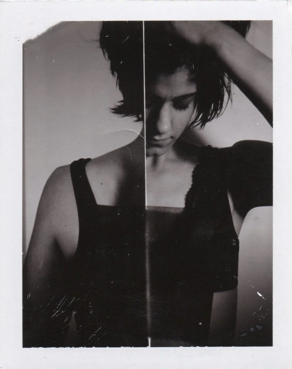 Black and white photo of a young woman with short, tousled hair, wearing a sleeveless top, with her head tilted downward and one hand resting on her head.