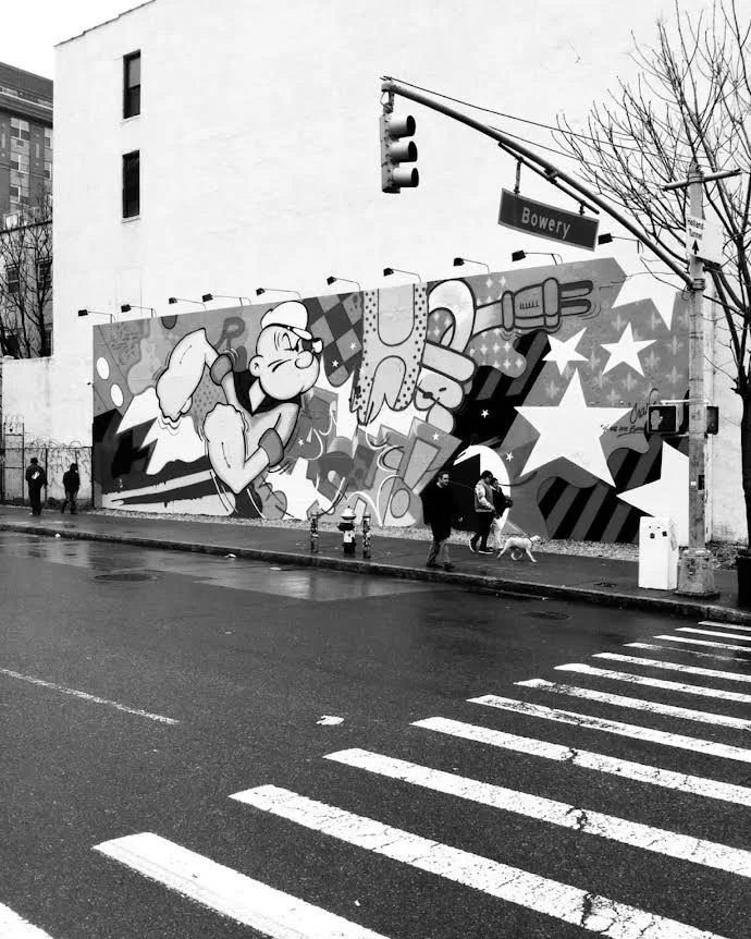A mural of a cartoon character in a baseball uniform with a baseball cap, pointing and smiling. The mural features stars and abstract shapes. It is painted on the side of a building on a city street corner, with pedestrians and a dog visible, and a s