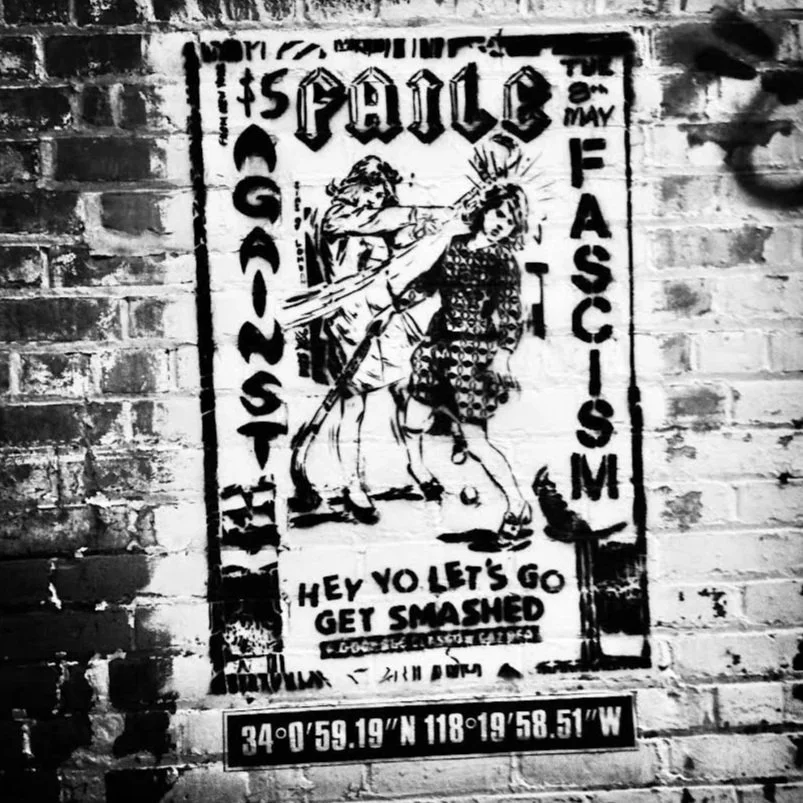 Street art poster on a brick wall depicting a fight scene with two women and bold text reading 'is PAULE', 'GAS LIGHT', and 'FASCISM'. Additional text says 'HEY VOLET'S GO GET SMASHED'. Coordinates at the bottom: 34°0'59.19"N 118°19'58.51"W'.