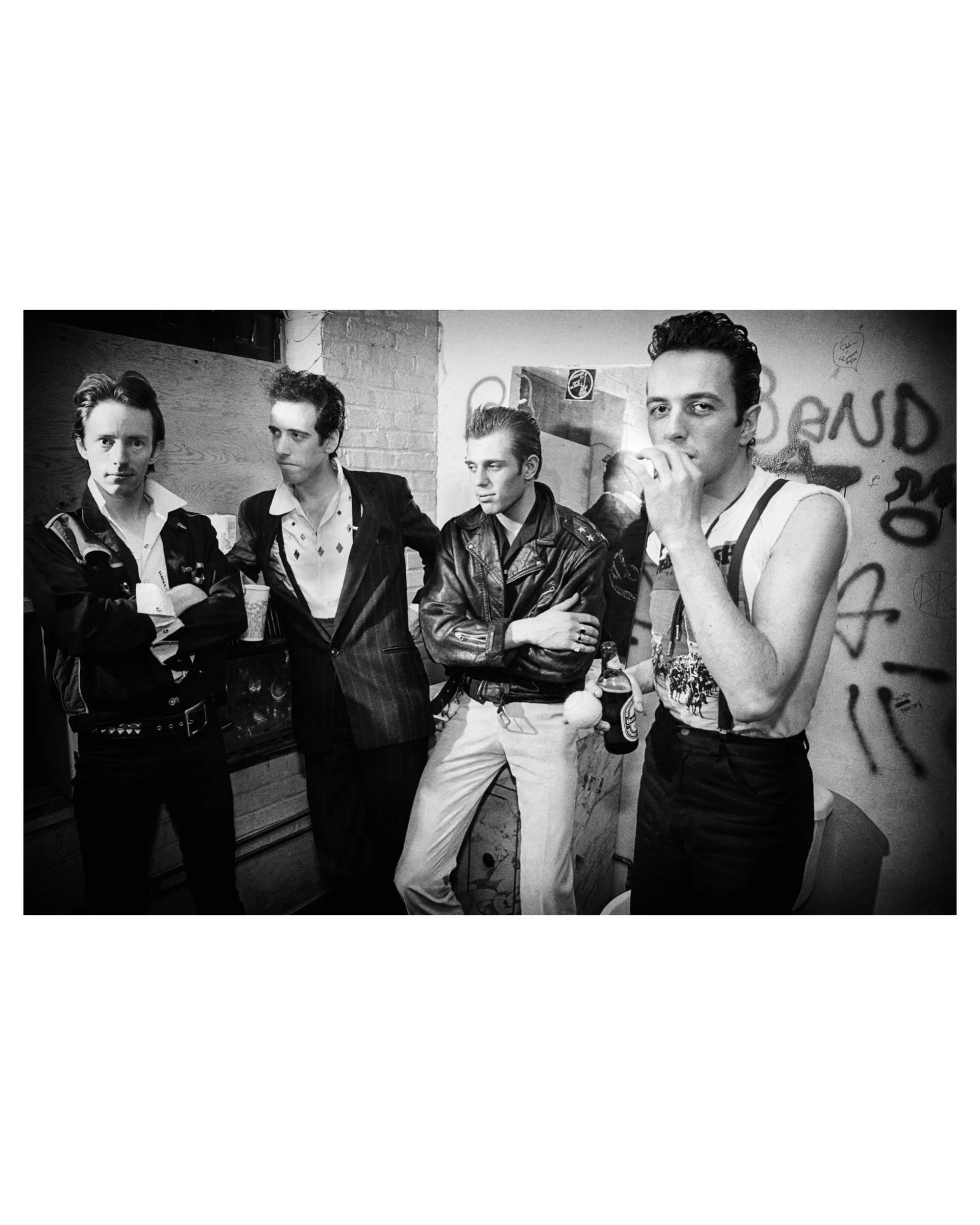 Black and white backstage portrait by Michael Grecco of the band The Clash, four members standing against a graffiti-covered wall.