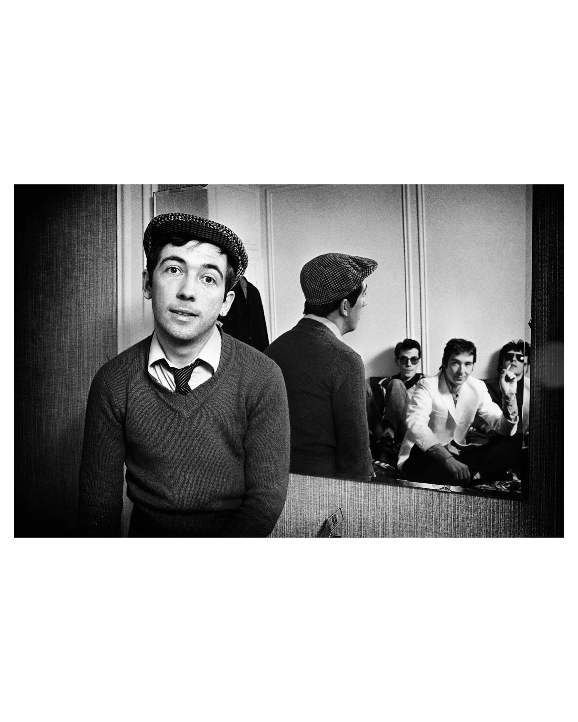 Black and white backstage photo by Michael Grecco of Pete Shelley in a flat cap and sweater, with Buzzcocks bandmates reflected in a mirror behind him.