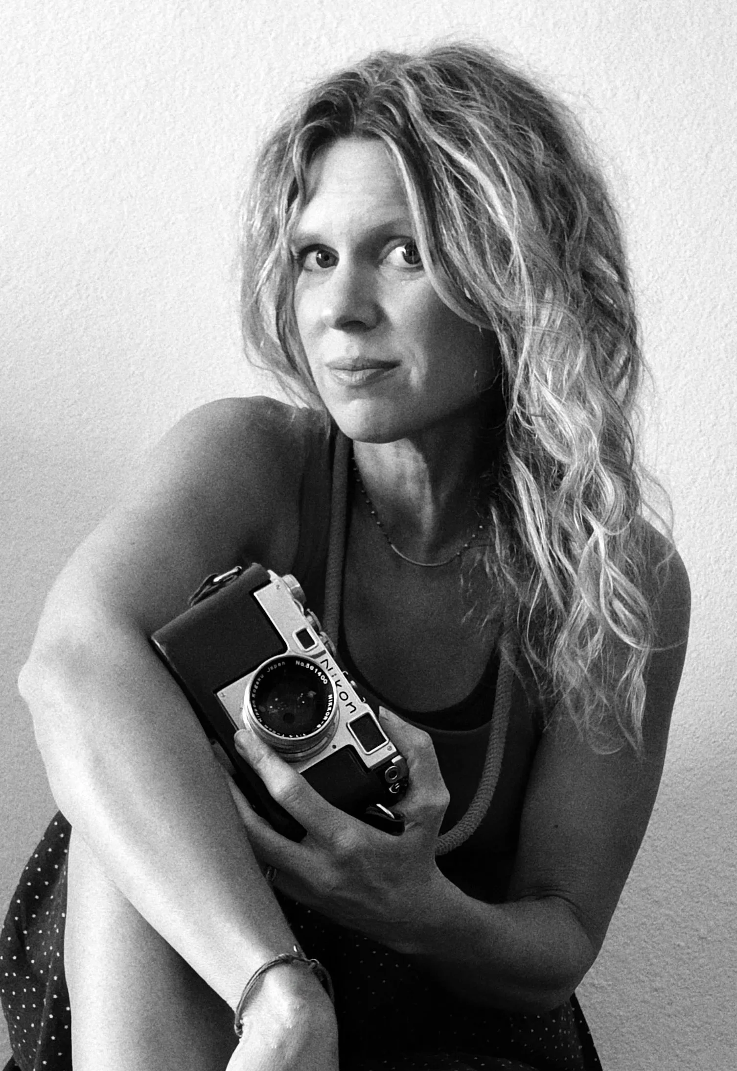 Black and white photo of photographer, Leigh Ann Edmonds. She is looking at the camera with a subtle smile. She is wearing a sleeveless top, a necklace, and a watch on her wrist. The background is plain wall.
