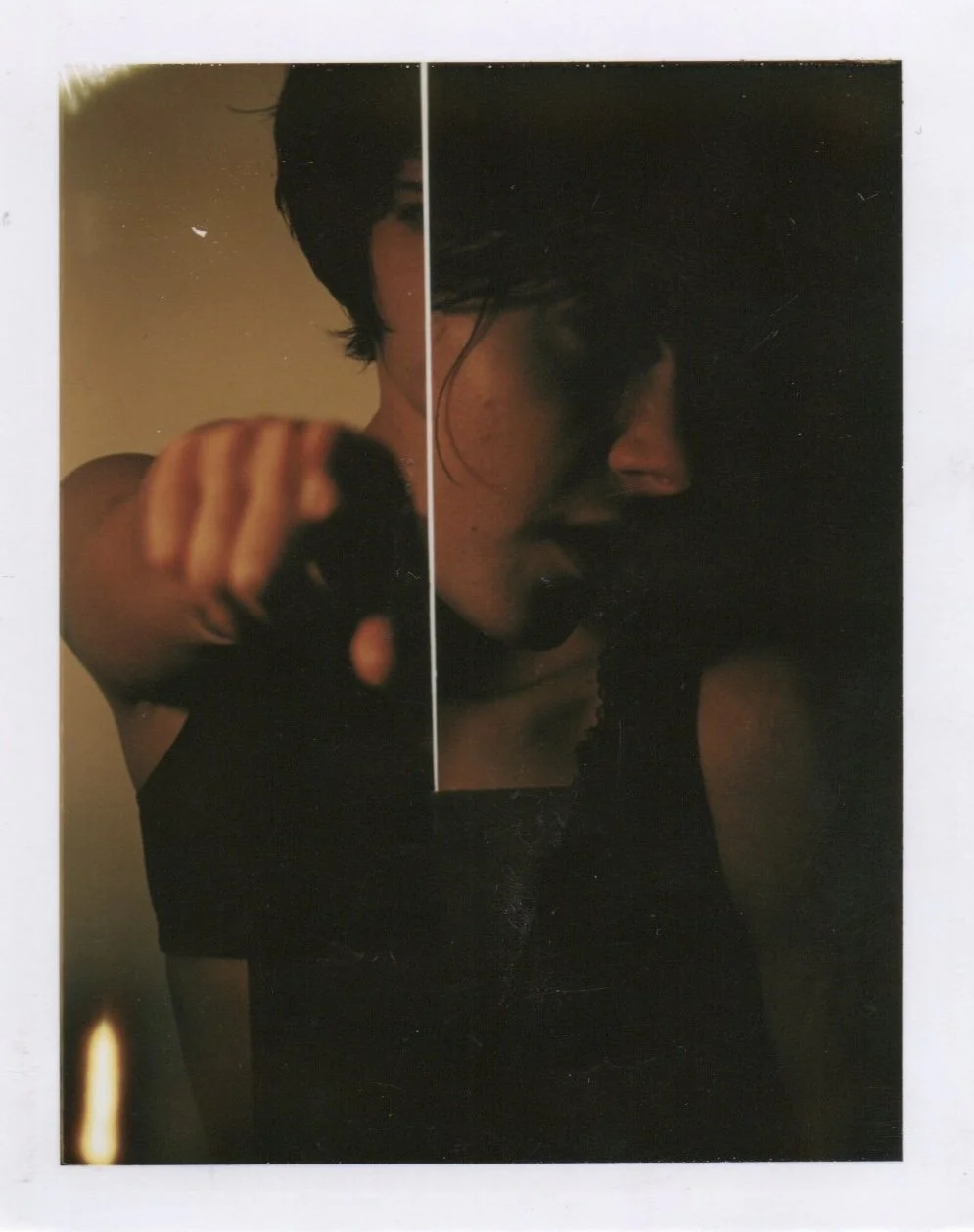 A person with dark hair wearing a black top, shot in low light, with half of their face obscured by a mirror or reflective surface, pointing directly at the camera.