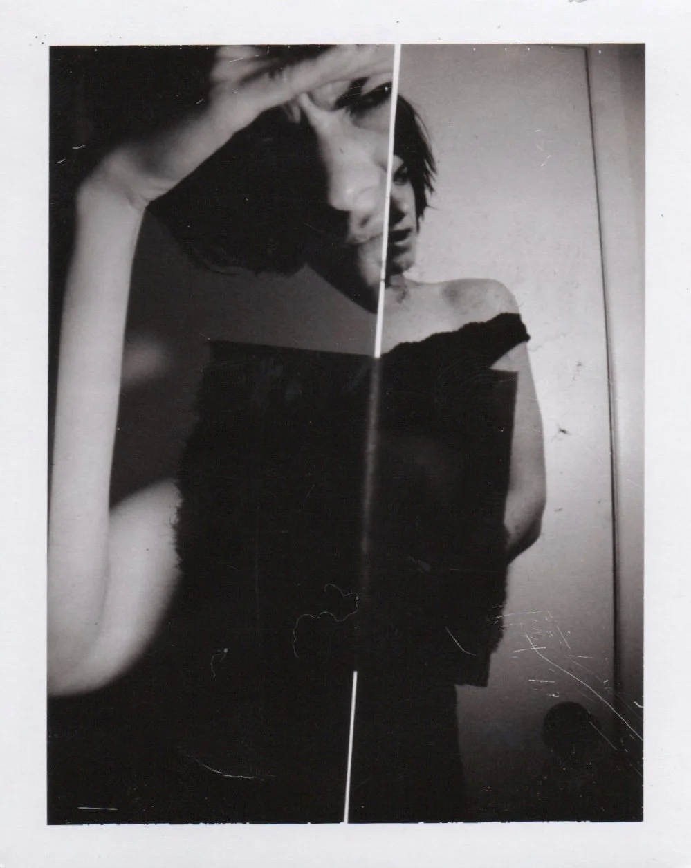 Black and white photo of a woman taking a selfie with her phone, holding it with her left hand. She has shoulder-length dark hair and is wearing a strapless top. The photo is mirrored and creased in the middle.