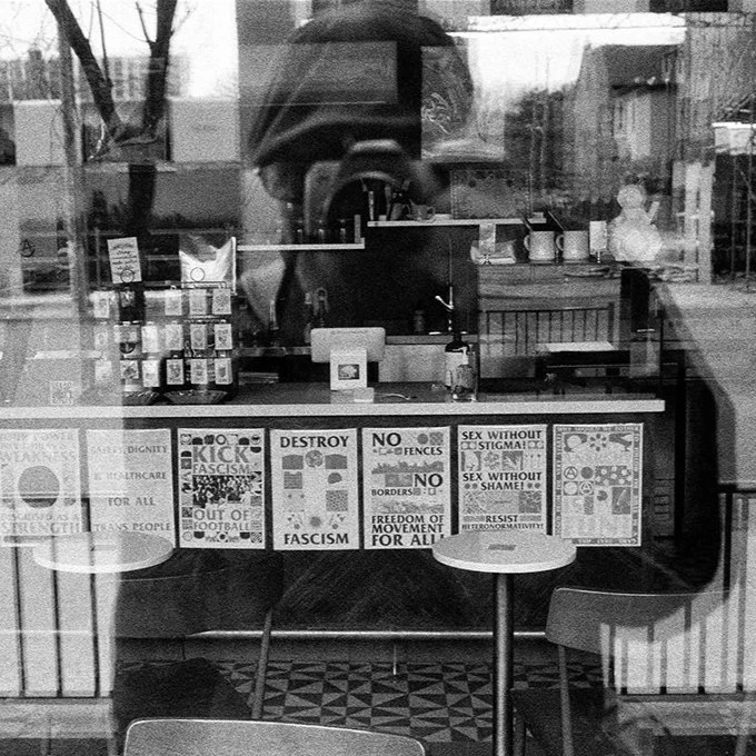 Reflection of Dave Green inside a cafe or shop, with posters and signs on the window, including messages about anti-fascism and social issues, and a counter with items on display.
