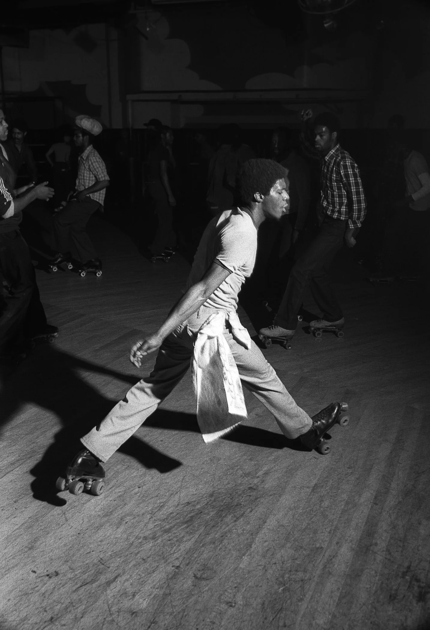 A man skating on roller skates in a large indoor space, wearing light-colored pants, a light shirt, and a jacket tied around his waist, with multiple people in the background from the collection "Empire Roller Disco' by Patrick D. Pagnano.