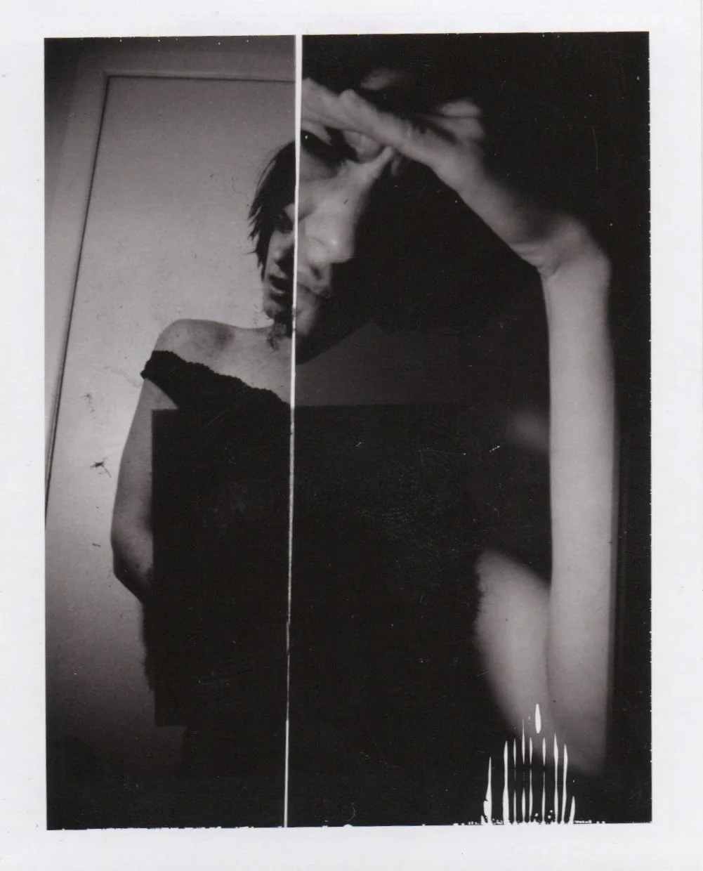 Black and white photograph of a woman taking a mirror selfie, holding her phone in her right hand to cover part of her face, with her dark hair framing her face, wearing an off-the-shoulder top, and standing in a room with plain walls and a door.