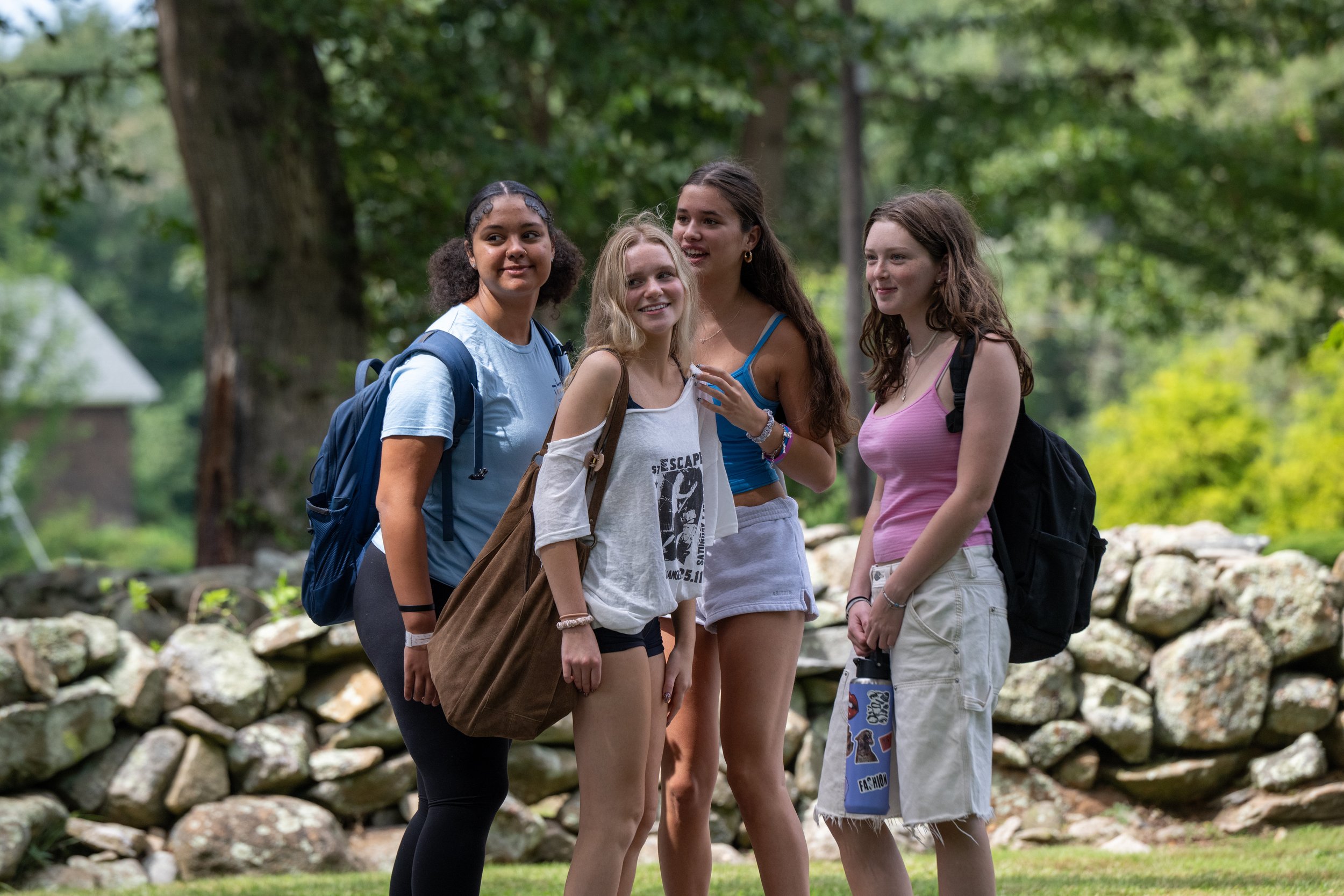 Four teenage girls standing outdoors in a wooded area, smiling and talking, with backpacks, near a stone wall.