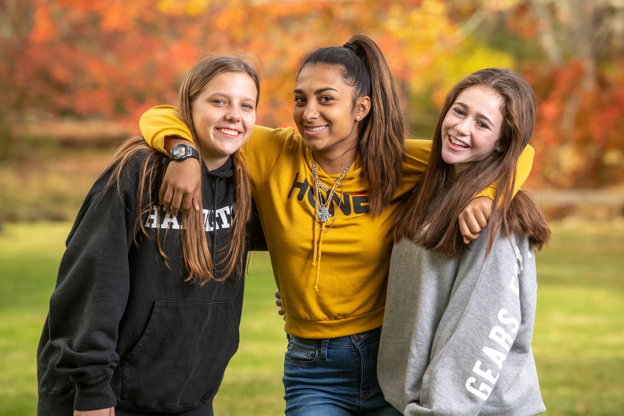 Three teenage girls outdoors, smiling and embracing, wearing casual hoodies, with autumn foliage in the background.
