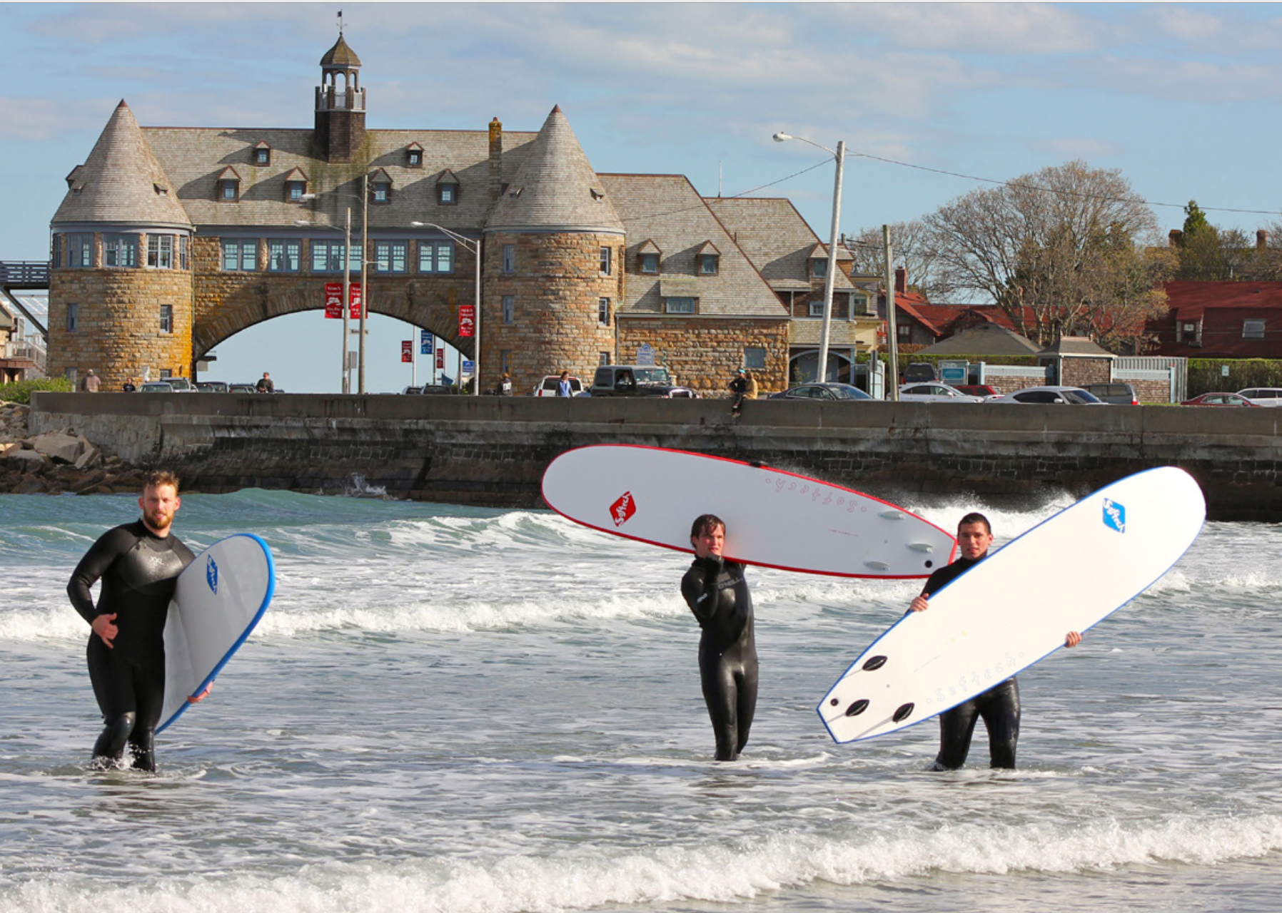 Three surfers in wetsuits standing in ocean water near a beach with a historic stone castle behind them