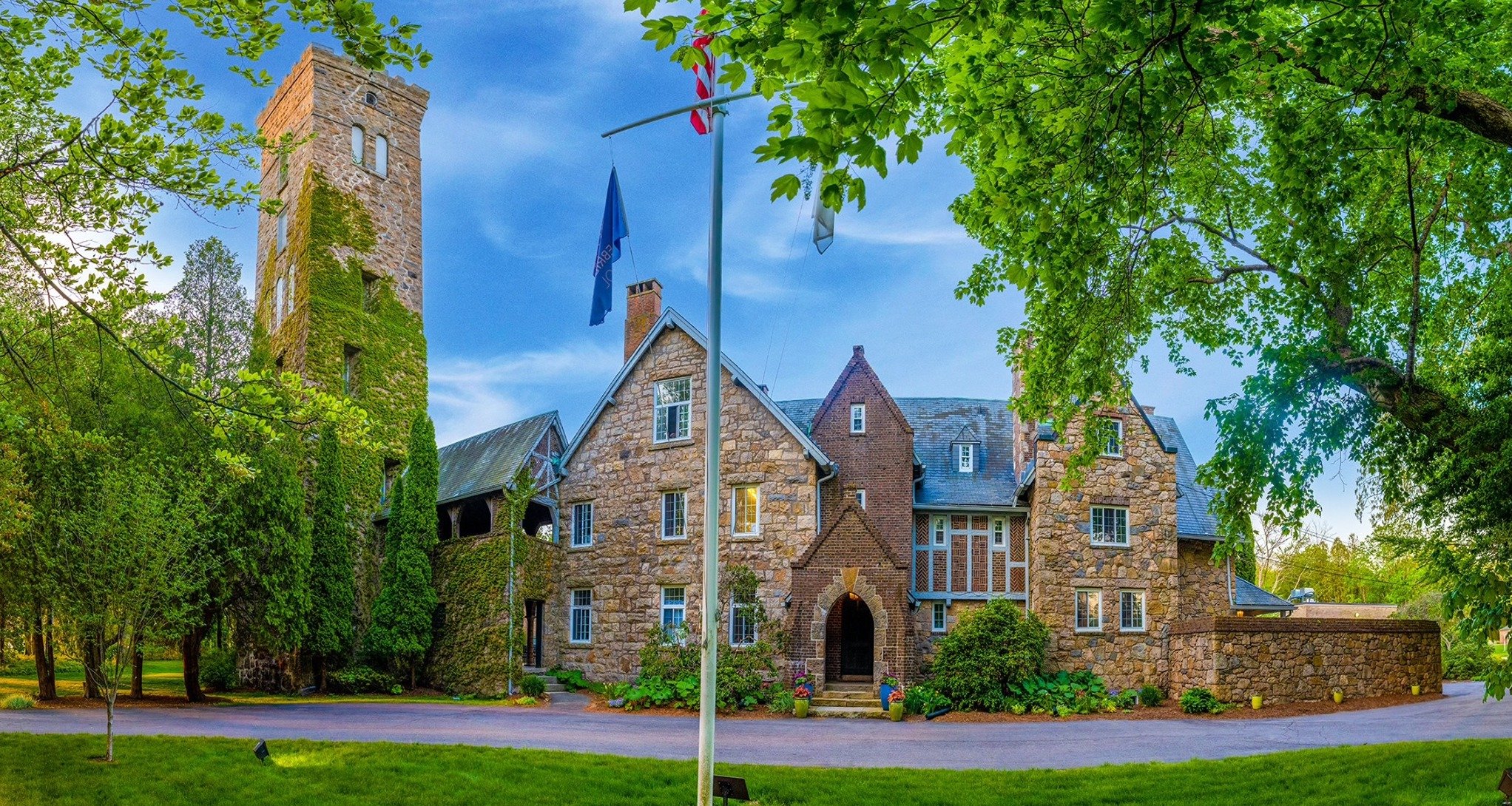 A large stone and brick house with pointed roofs, surrounded by green trees and a lawn, with flags flying on a flagpole in front.