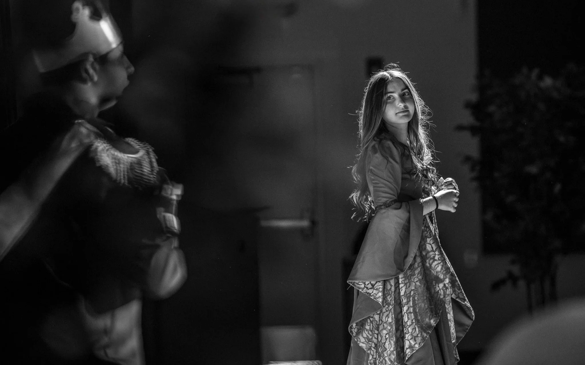 A young woman with long hair looking over her shoulder in a black-and-white photo, standing near a Christmas tree, holding a glass, with two children in cowboy hats nearby.