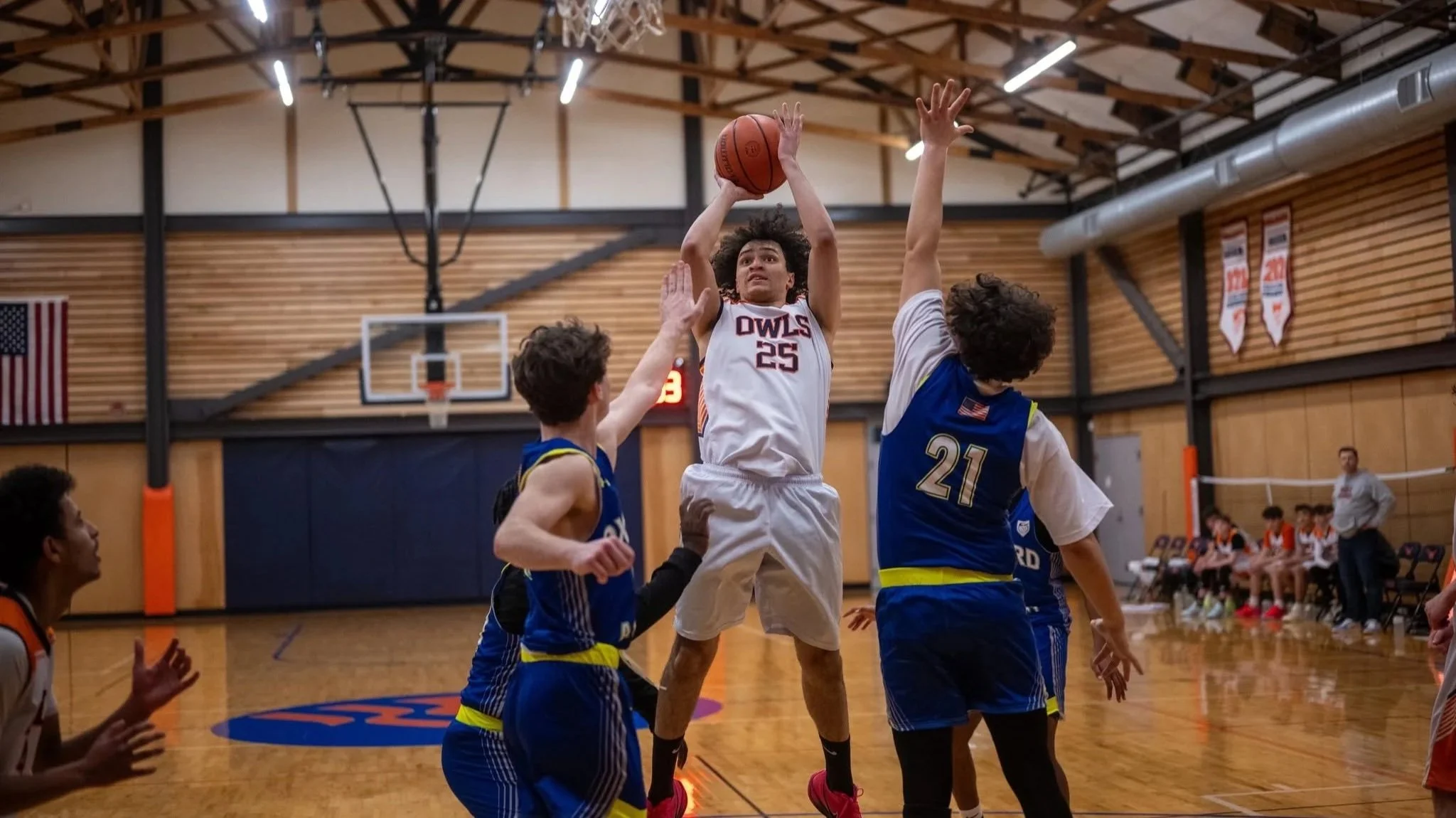 A basketball game with players jumping for the ball inside a gymnasium. The player in white, wearing jersey number 25, is attempting a shot while others in blue jerseys try to block him. The gym has wooden walls, a basketball hoop, and an American fl