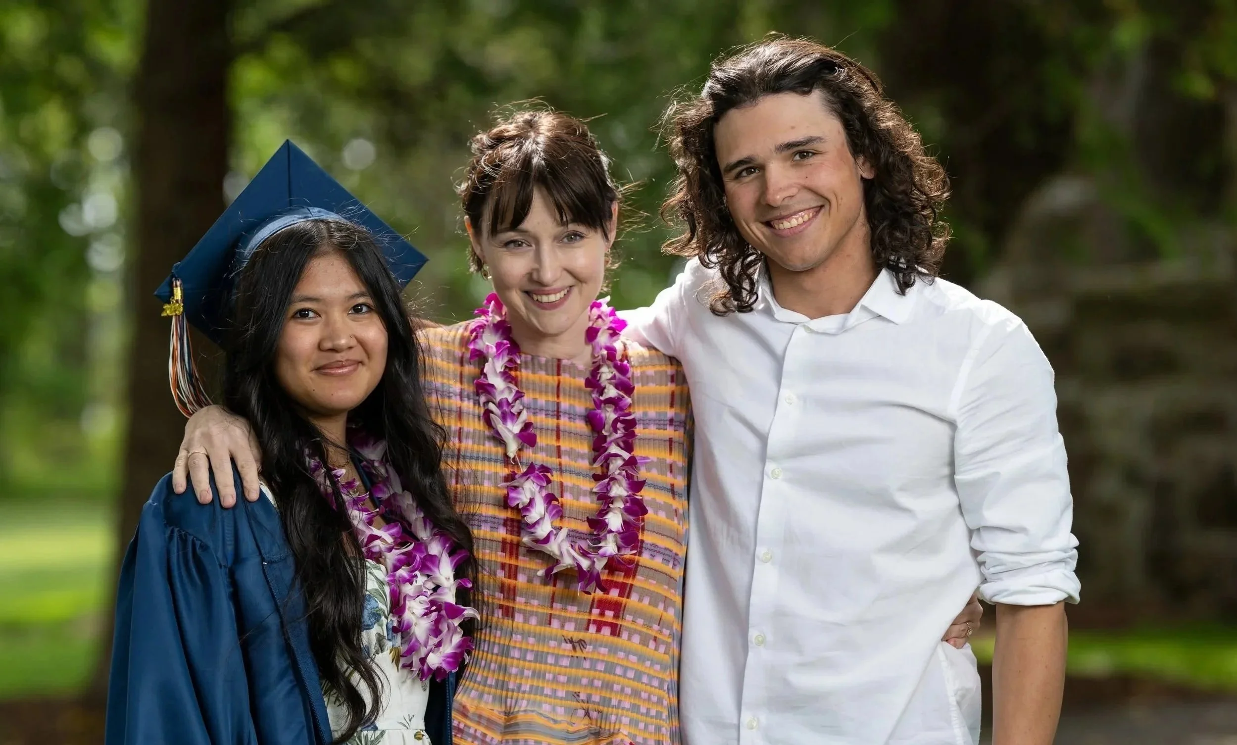 Three people smiling outdoors, one girl in a graduation gown and cap, another woman wearing a lei, and a man in a white shirt, with a blurred green forest background.