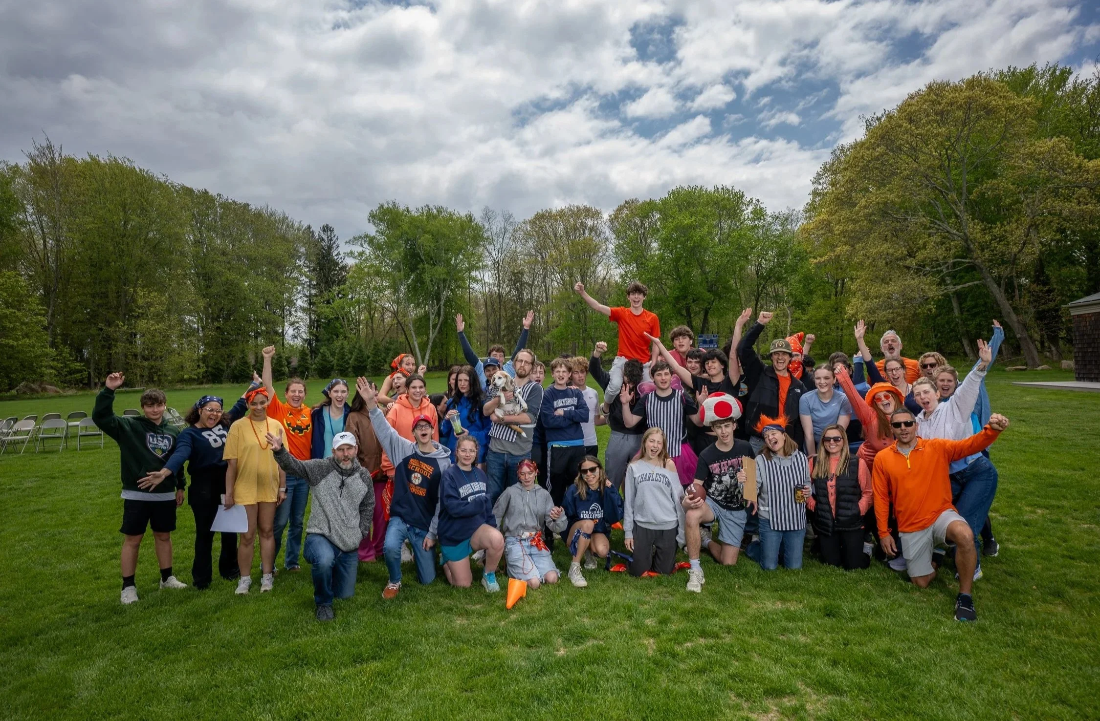 A large group of people gathered outdoors on a grassy field, celebrating, with many raising their arms, some wearing Halloween costumes, and a few holding drinks. Trees and a cloudy sky are in the background.