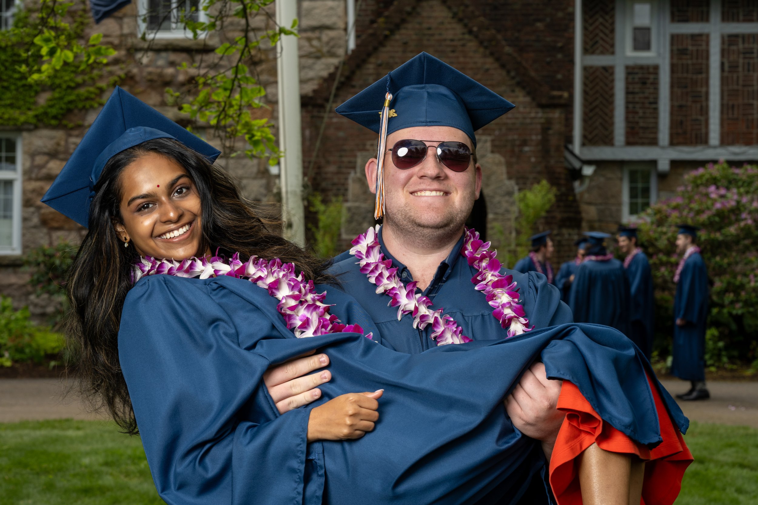Grad graduate in blue cap and gown being carried by a smiling woman in a similar blue cap and gown during outdoor graduation ceremony. Both are wearing leis, with other graduates visible in the background.