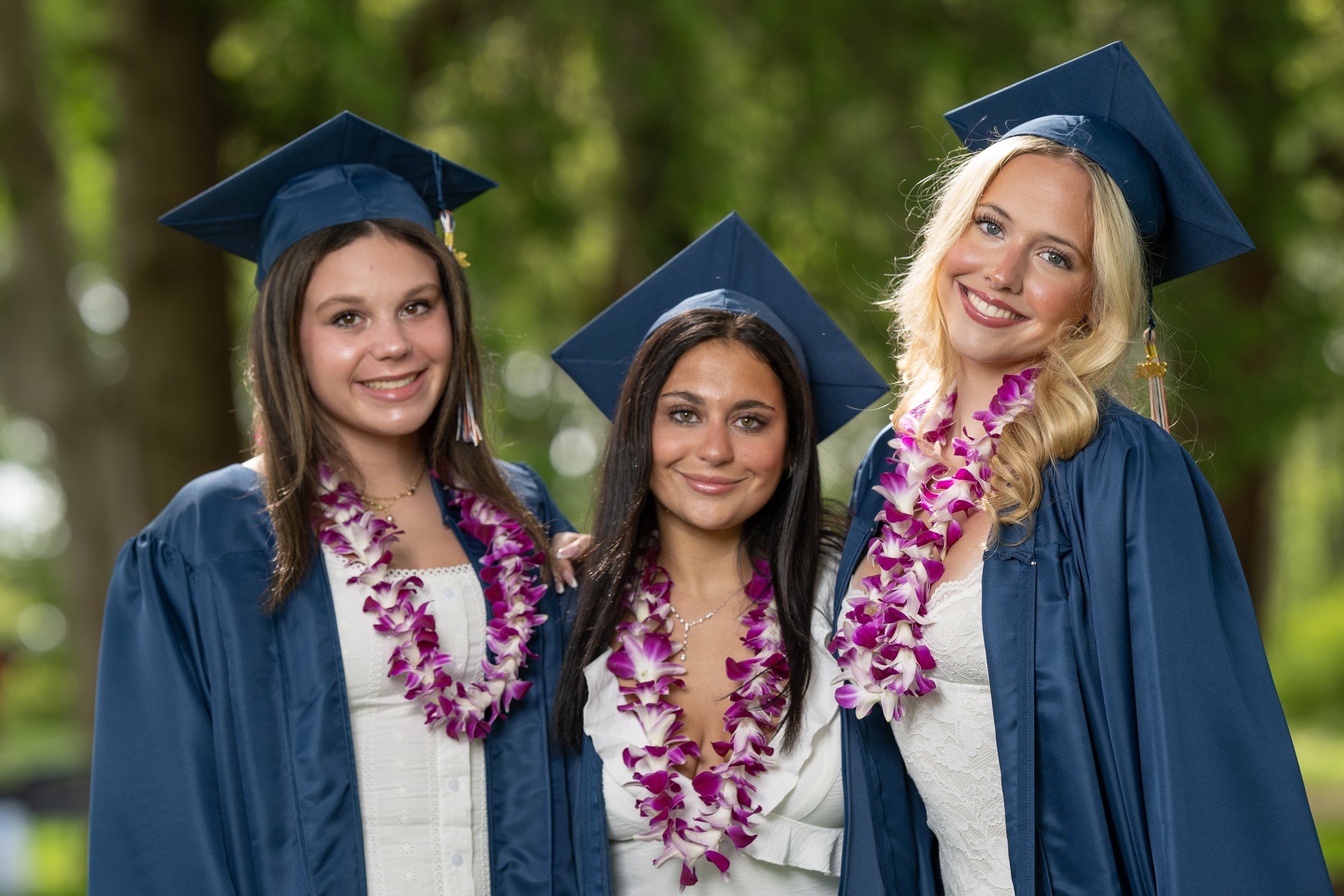 Group of four young women in graduation caps and gowns with leis, smiling outdoors with a blurred green background.