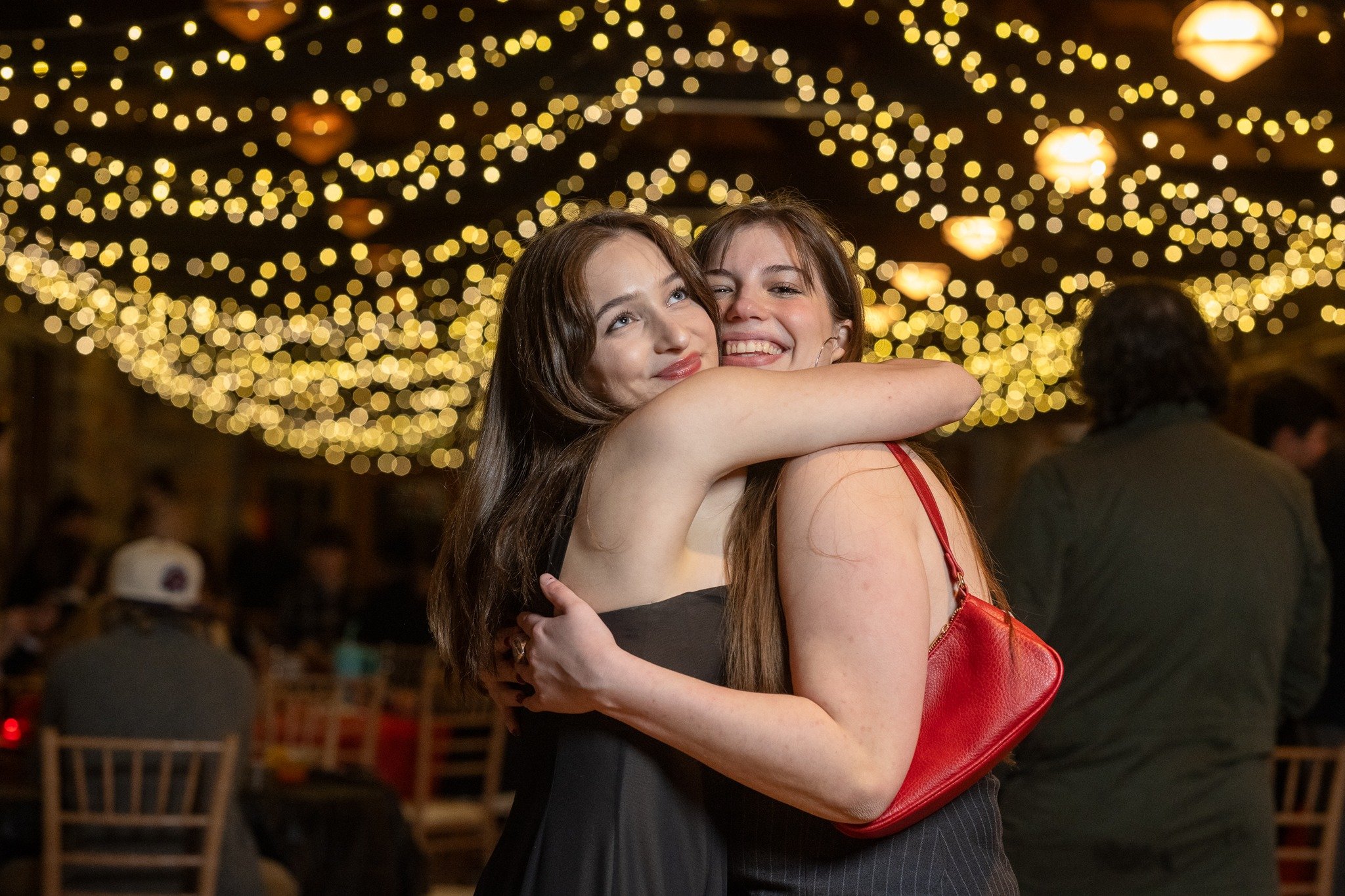 Two women hugging and smiling at a festive party with string lights and blurred guests in the background.