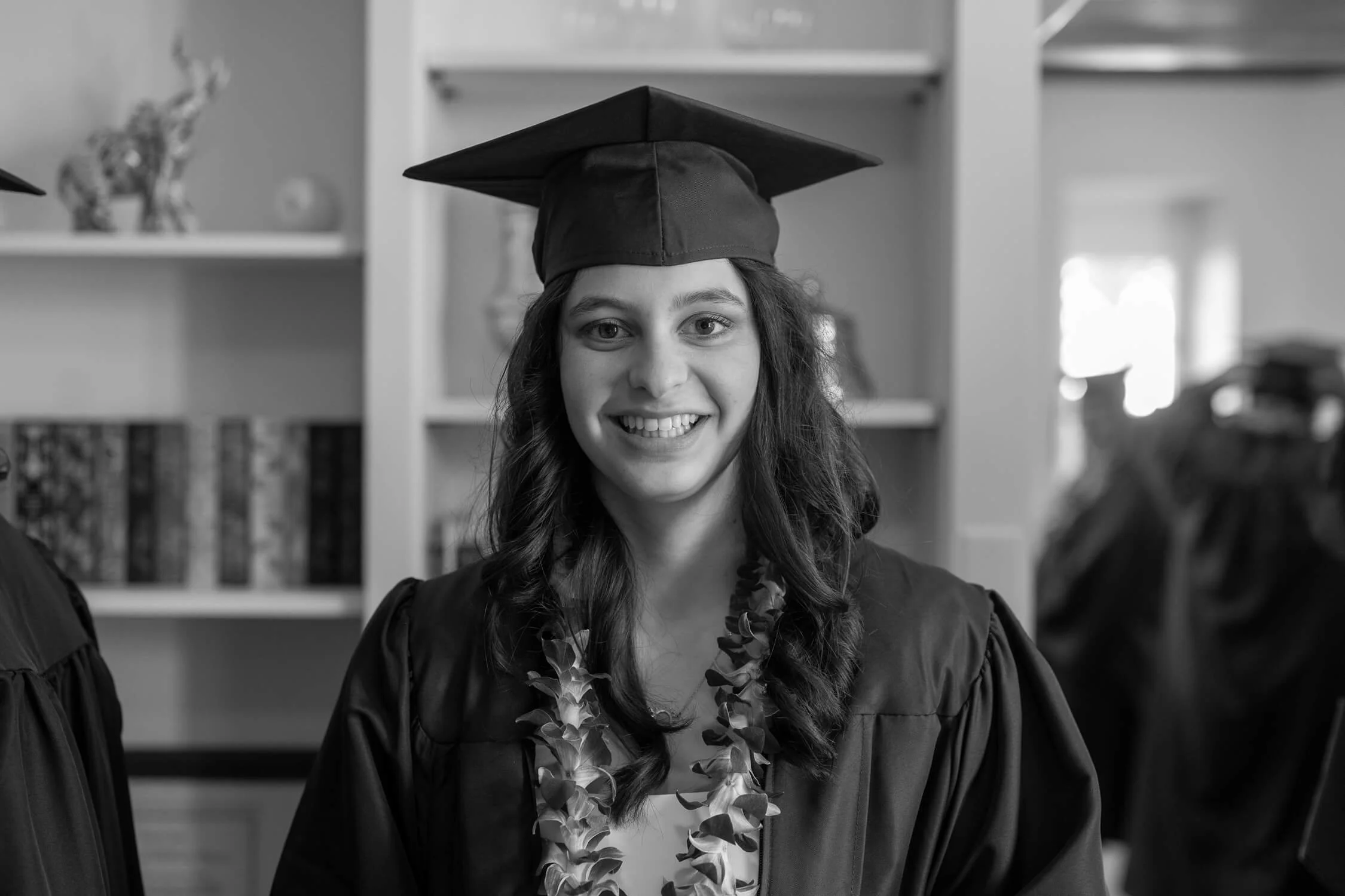 A young woman in graduation gown and cap smiling at the camera, wearing a lei, in an indoor setting.