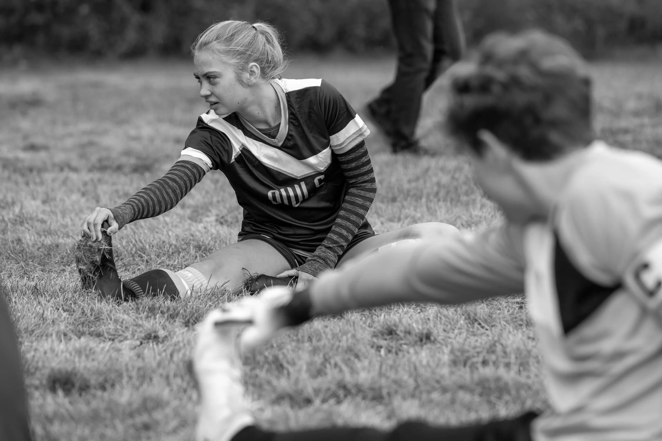 A young female athlete in a soccer uniform sitting on the grass, stretching or recovering after a game or practice, with a focused expression, while another player is partially visible in the foreground.