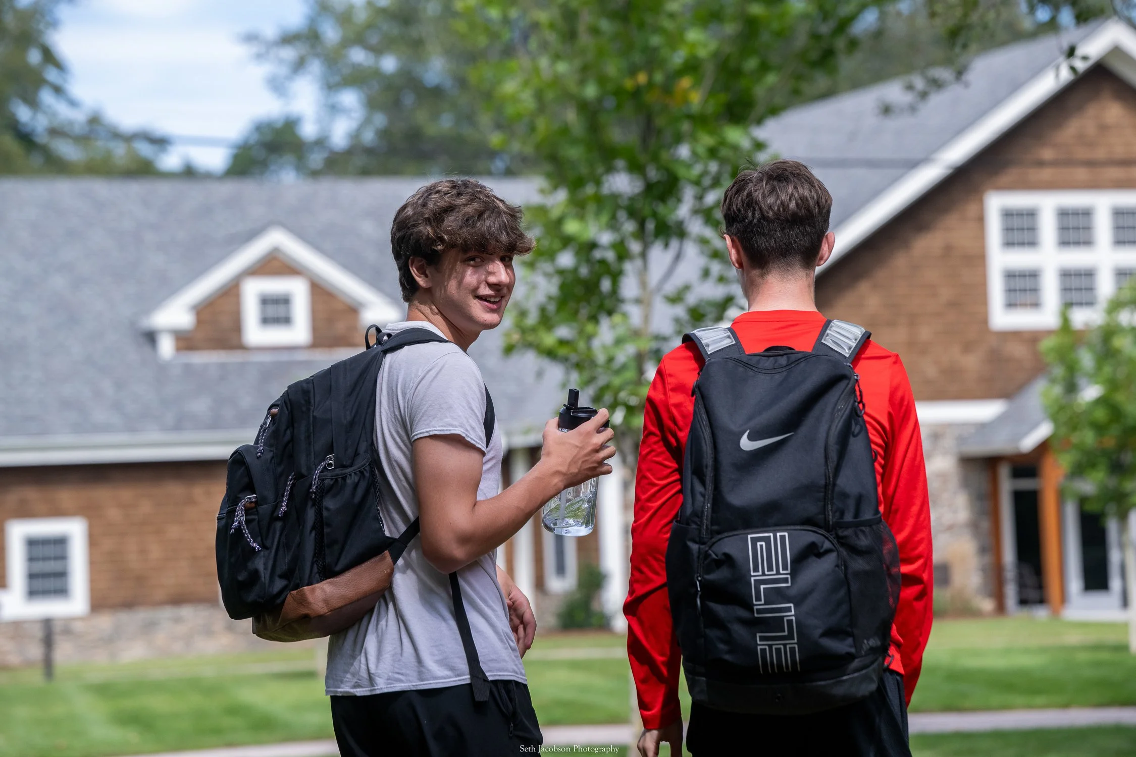Two young men with backpacks talking outdoors in front of a house with a gray roof and brown siding, surrounded by green trees.
