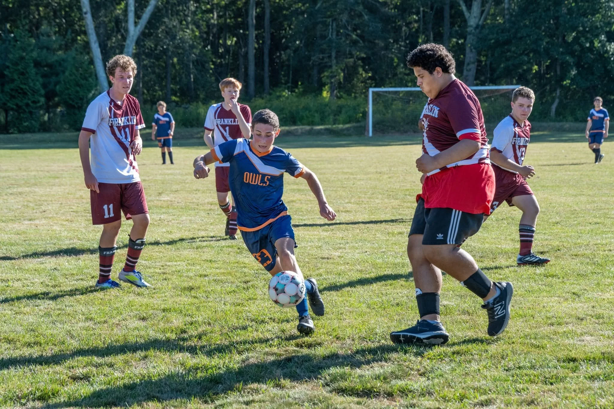 A group of boys playing soccer on a grassy field, surrounded by trees, with some wearing maroon and white uniforms and others in blue and orange uniforms.