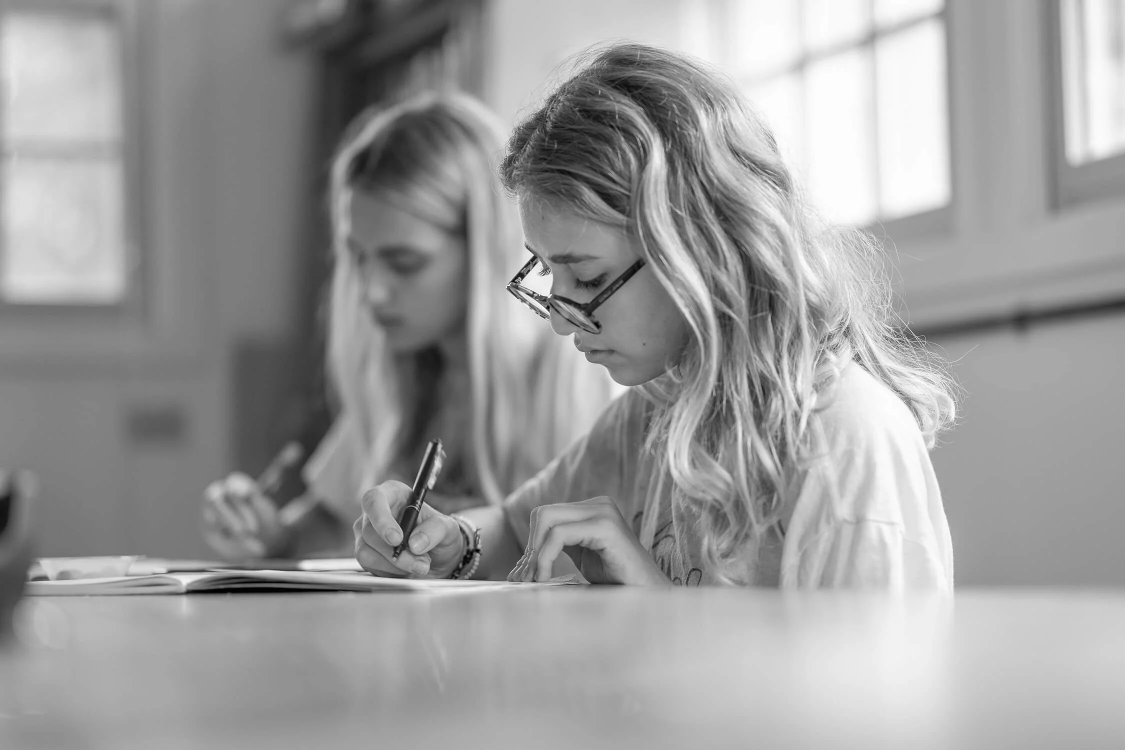Two young women with glasses are sitting at a table, writing on paper in a classroom with windows in the background.