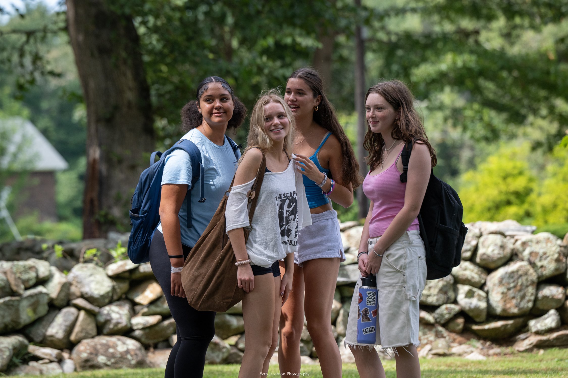 Four young women sitting on green grass outside, laughing and talking with each other, with a building and trees in the background.