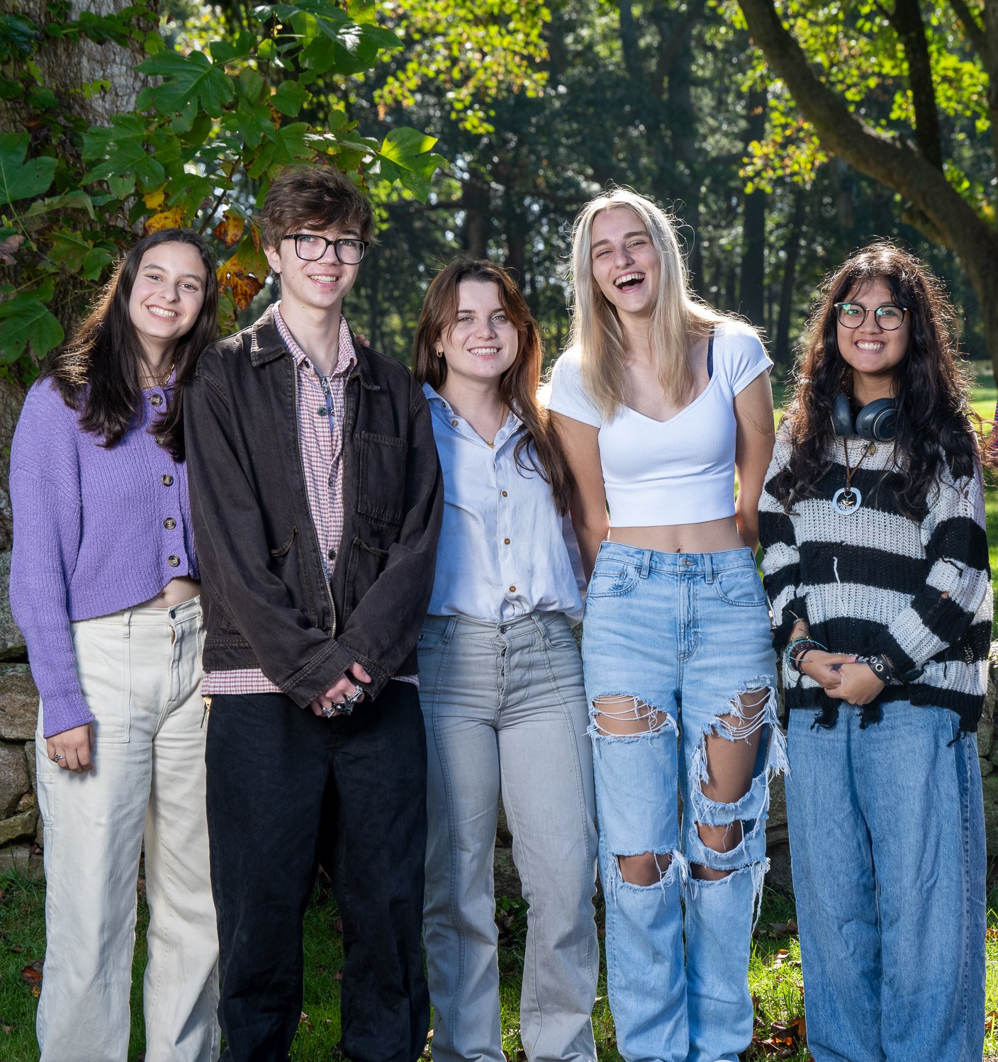 Group of five young women standing outdoors in a park, smiling and laughing, with trees and sunlight in the background.