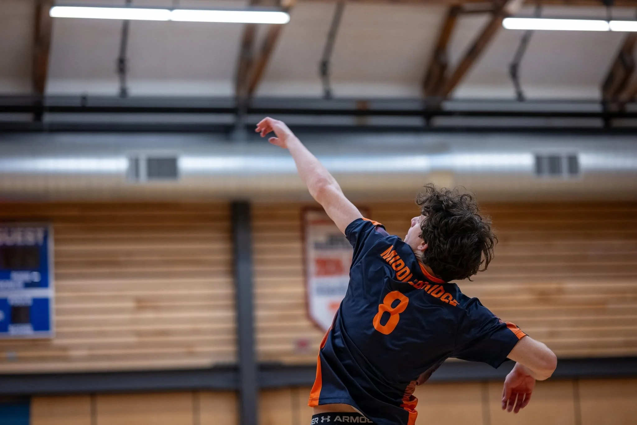 A young boy in a dark sports uniform with orange accents and the number 8 on his back playing a volleyball game indoors. The boy's arm is extended upward, likely hitting or reaching for the ball, in a gymnasium with a wooden ceiling and walls.