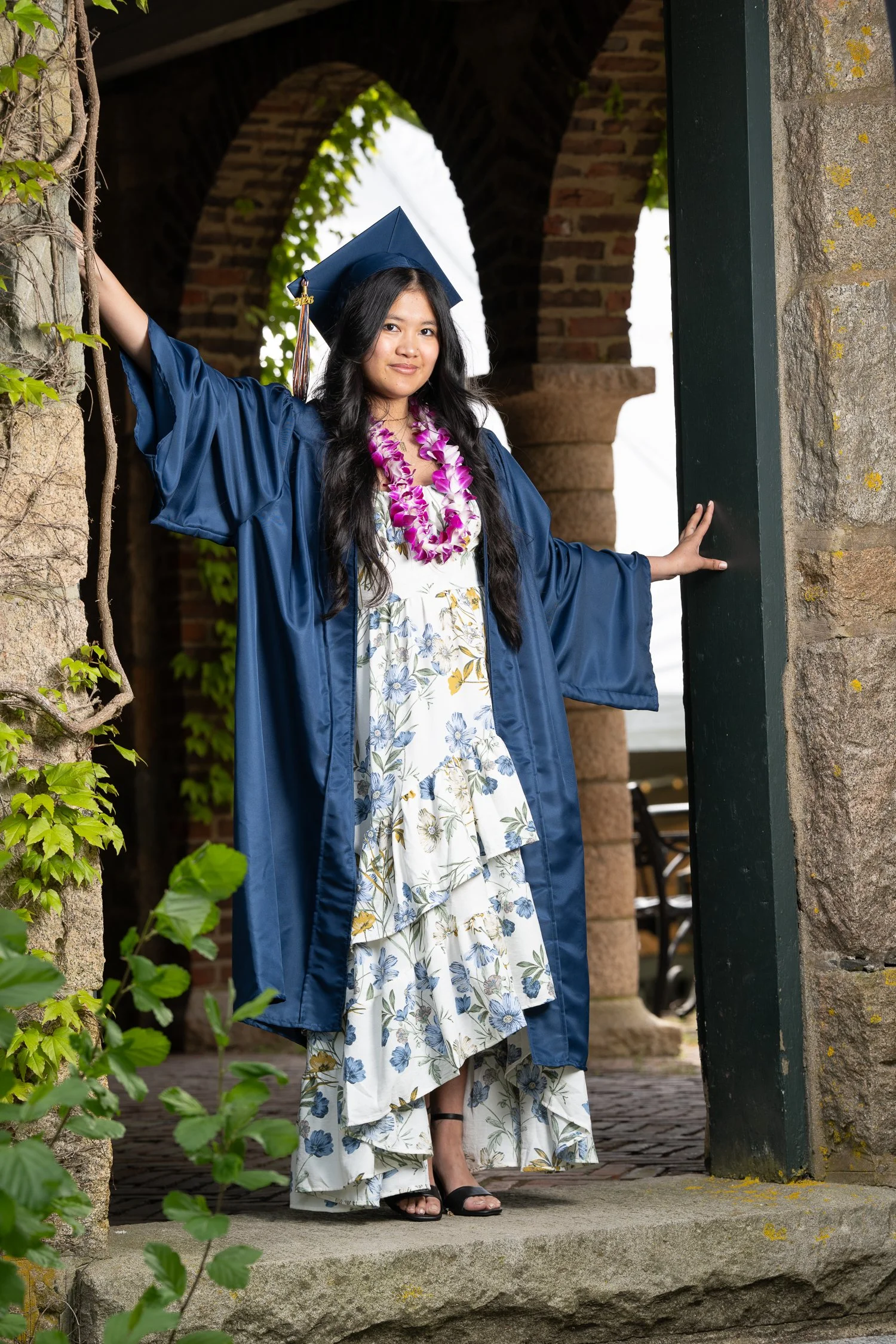 A young woman in a graduation cap and gown standing outside near a brick archway, wearing a floral dress and lei, posing with one hand on a stone wall.