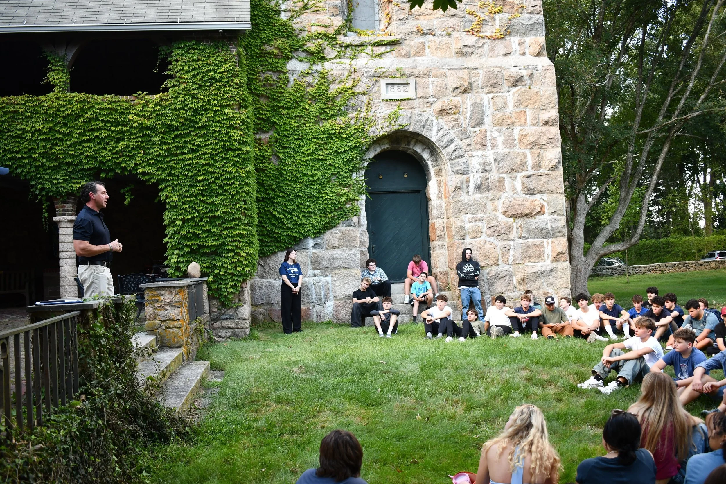 A group of children sitting on the grass, watching a man speaking in front of an outdoor stone building covered in green vines, with several adults standing nearby.