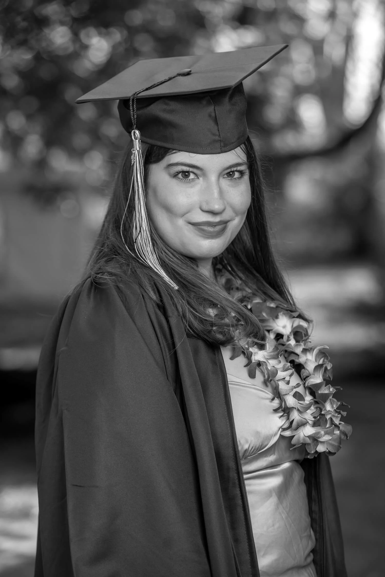 Black and white photo of a young woman in graduation cap and gown, smiling at the camera, with a floral lei around her neck, outdoors background.