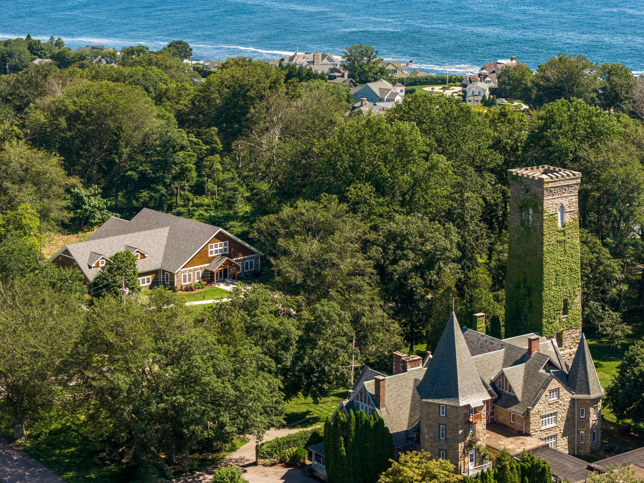 Aerial view of a neighborhood near the coast with several large houses, a stone tower, and dense green trees, overlooking the ocean on a sunny day.