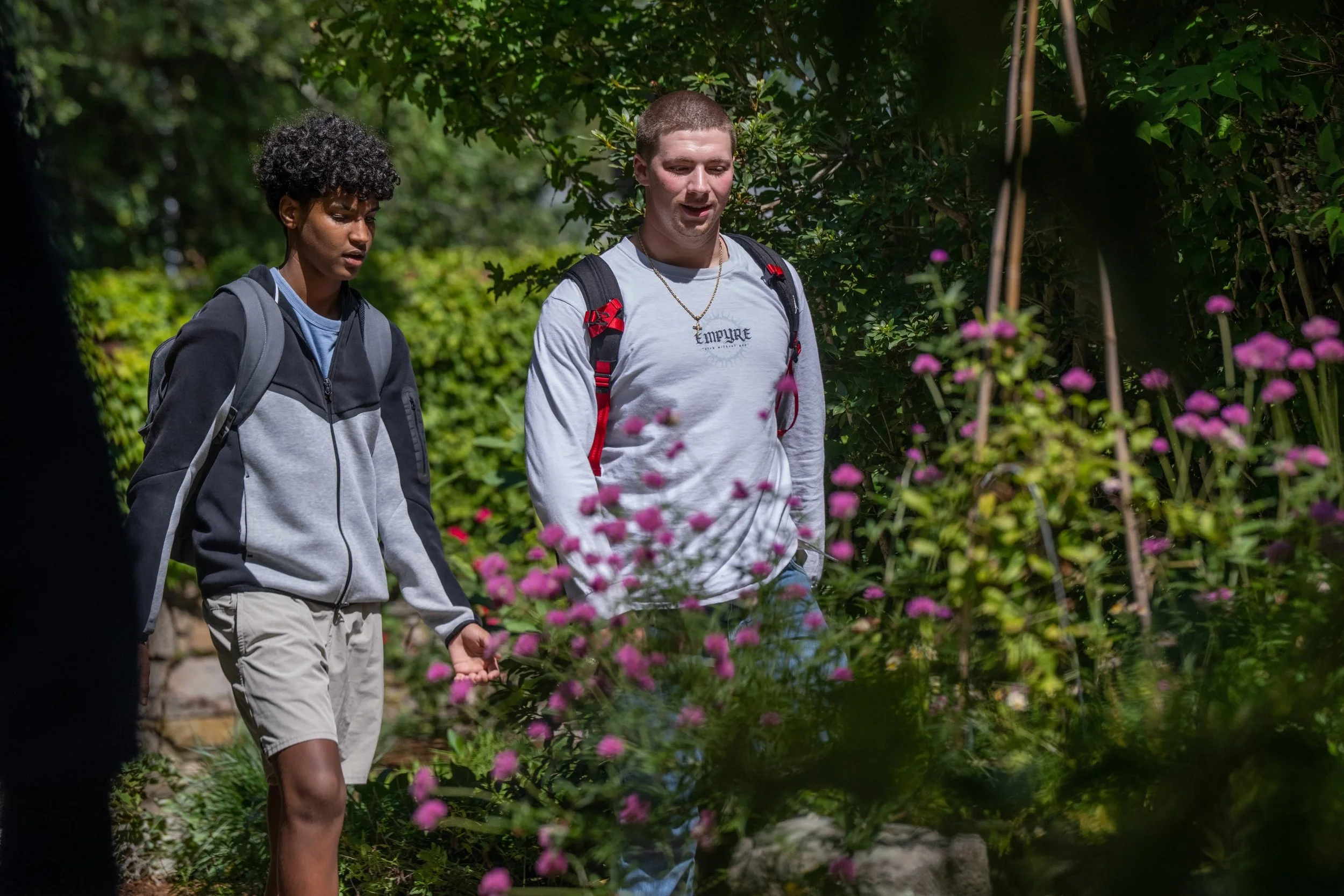 Two young men walking outdoors on a sunny day among green trees and pink flowers, one wearing a gray hoodie and the other wearing a light gray sweatshirt with a backpack.