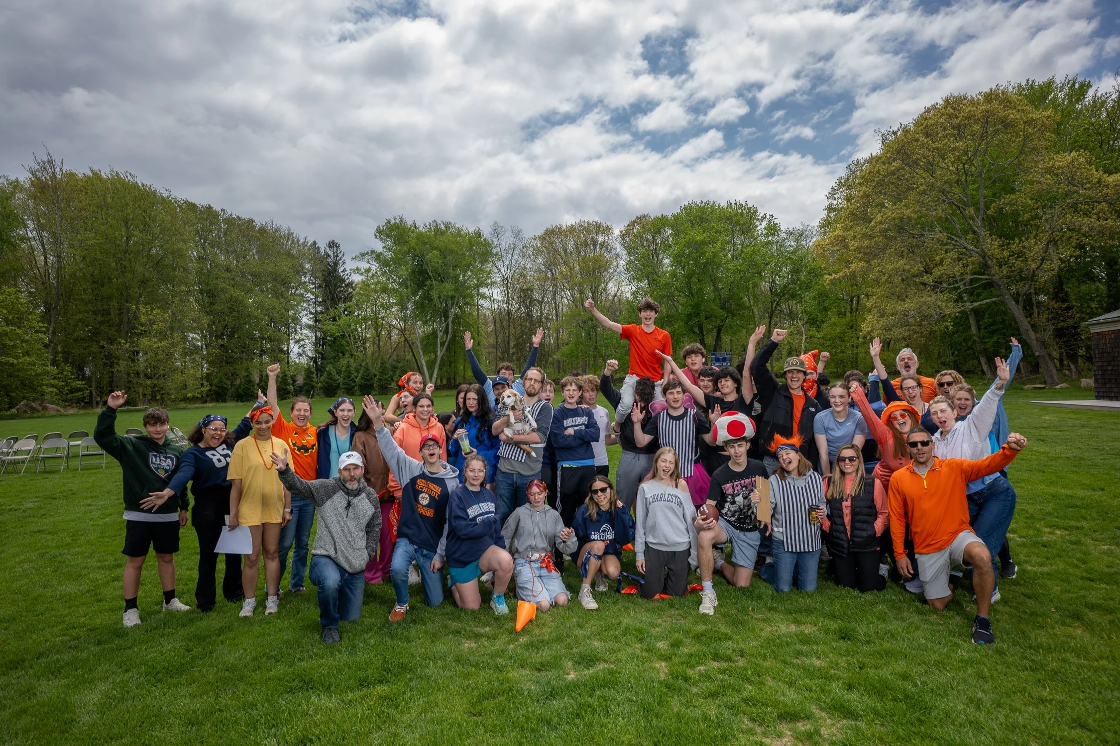 Group of people celebrating outdoors on a grassy field, with many raising their arms and smiling, dressed in colorful casual clothing, some wearing Halloween-themed accessories like a pumpkin face and a mushroom hat, surrounded by green trees under a