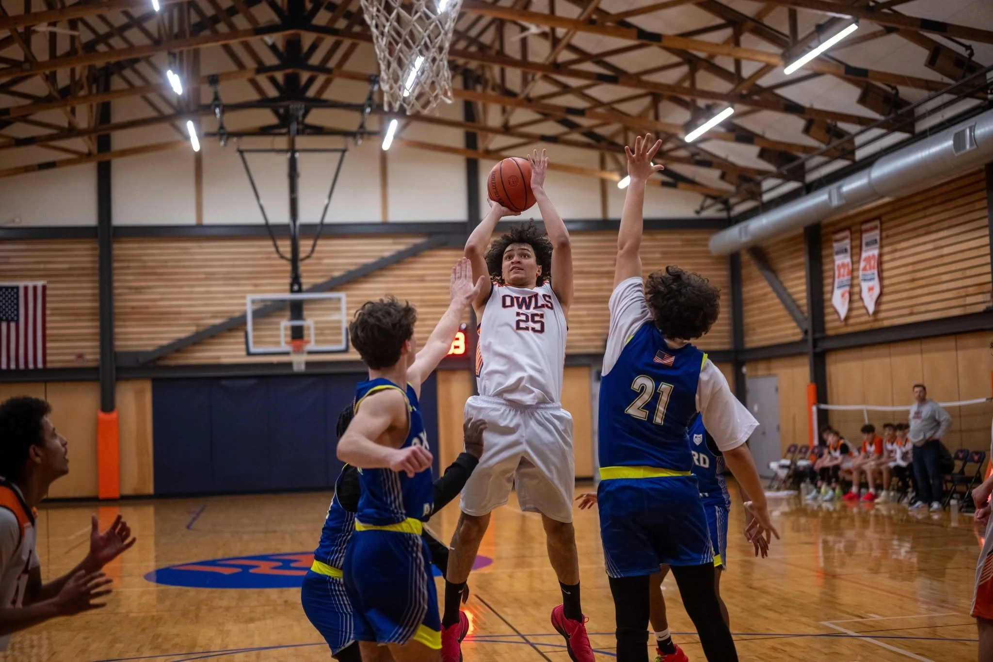 A basketball player wearing a white and orange uniform with the number 25 jumps to shoot the ball while being guarded by two opponents wearing blue and yellow uniforms inside a basketball gymnasium.