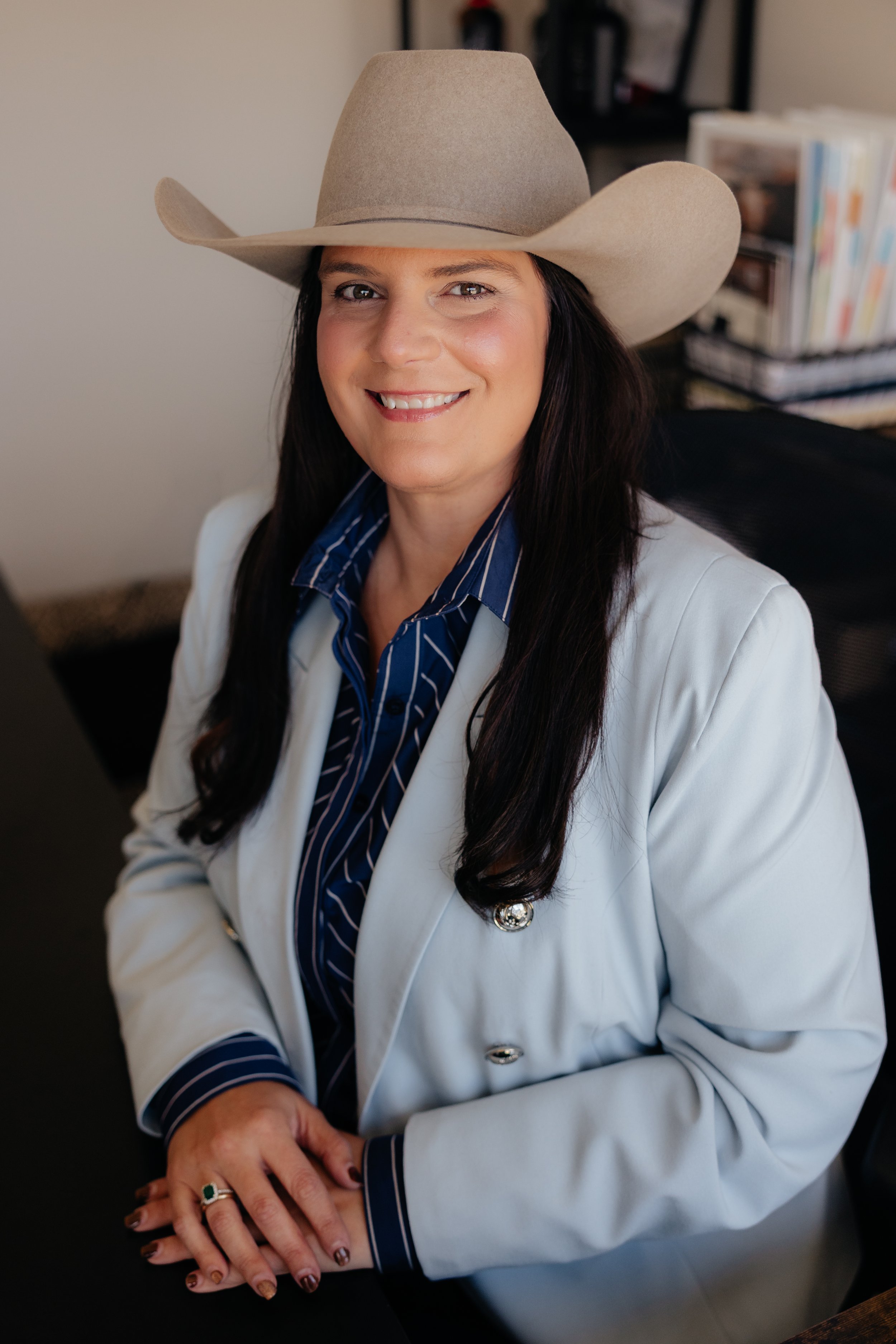 A woman smiling, wearing a white cowboy hat, a green blazer, and an off-white blouse, seated indoors.