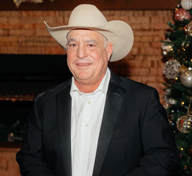 A man wearing a black cowboy hat, white shirt, and dark jacket, smiling indoors.