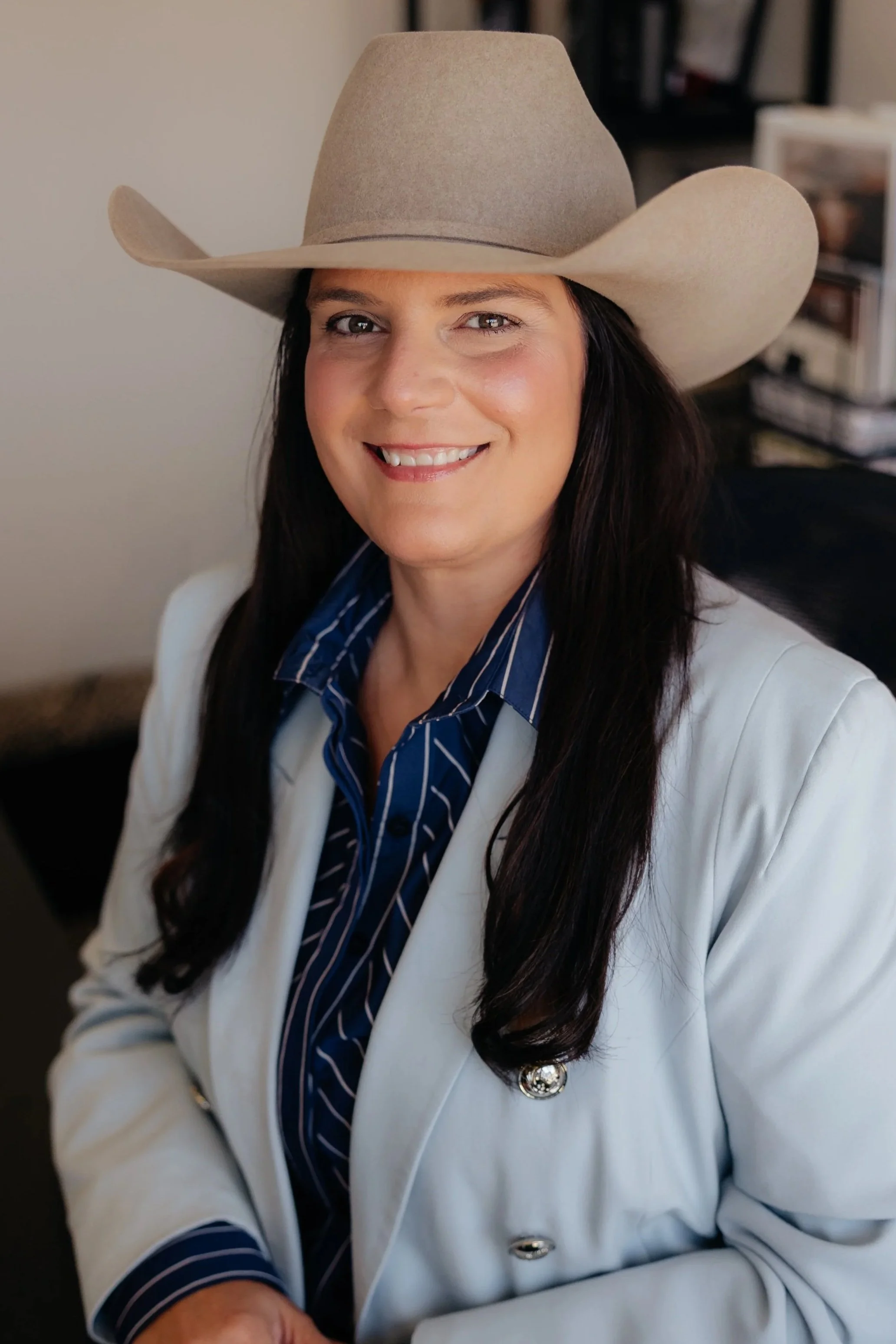 A woman smiling, wearing a white cowboy hat, a green blazer, and an off-white blouse, seated indoors.