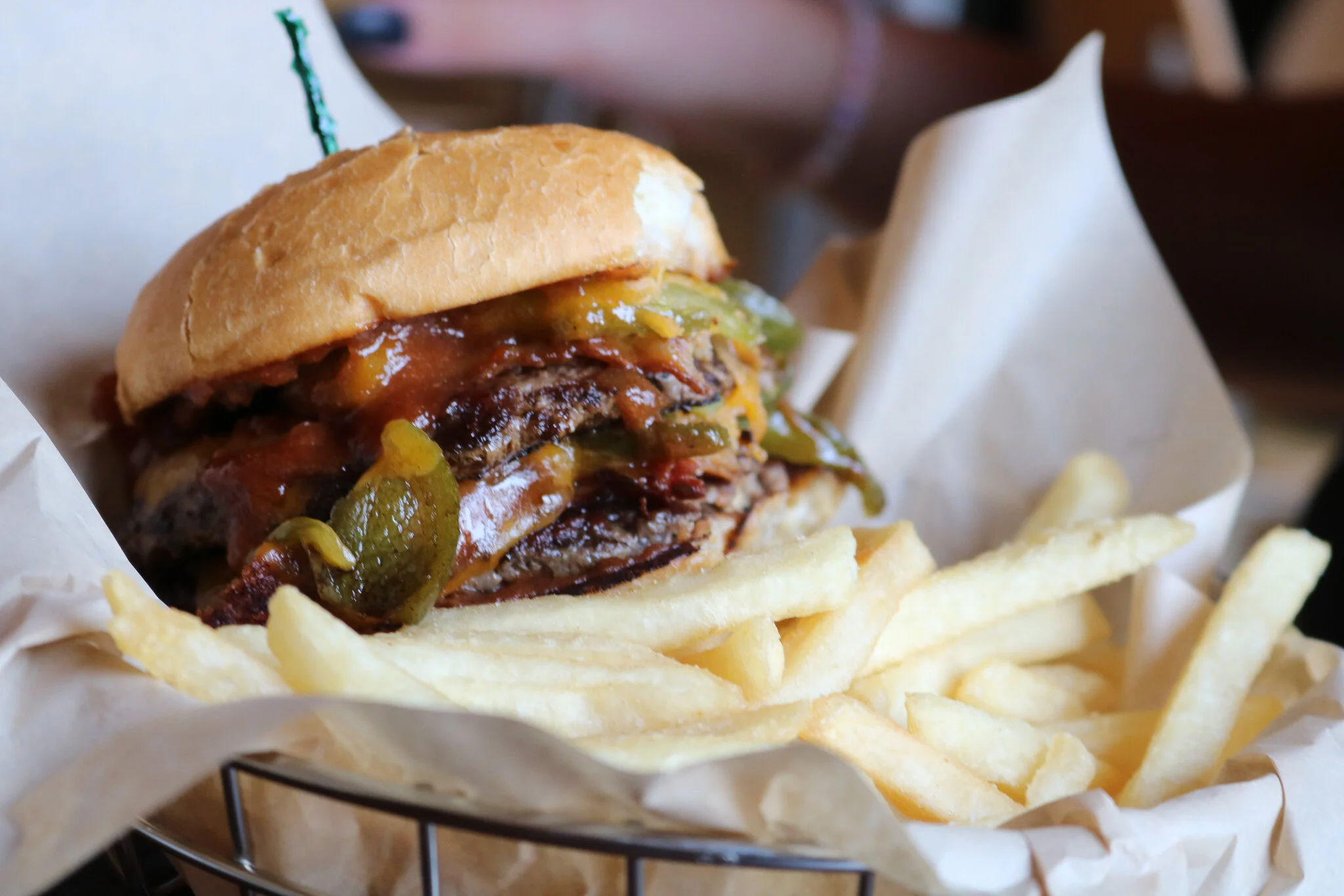 Close-up of a cheeseburger with lettuce, tomato, grilled onion, and a bun, served with a side of French fries in a metal basket with paper.