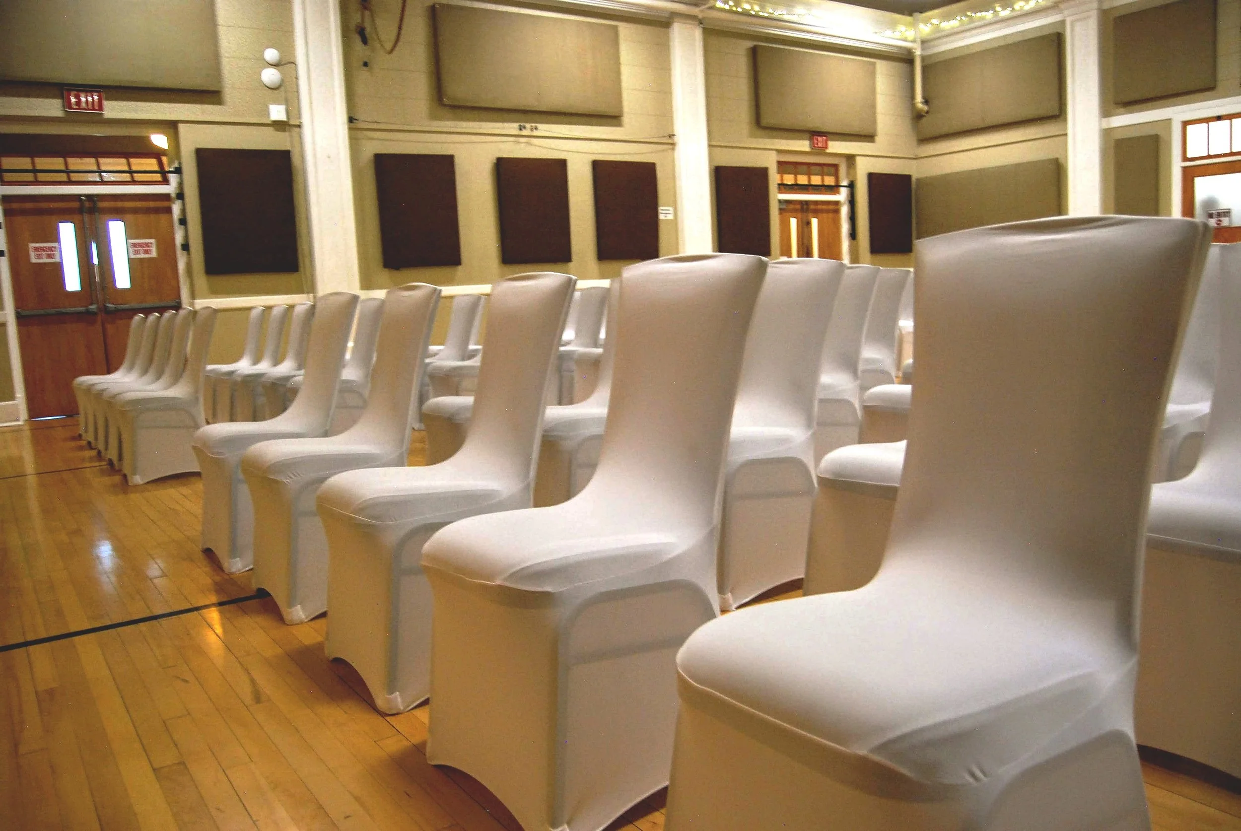 Empty white chairs arranged in rows inside a large room with wooden flooring and acoustic panels on the walls.