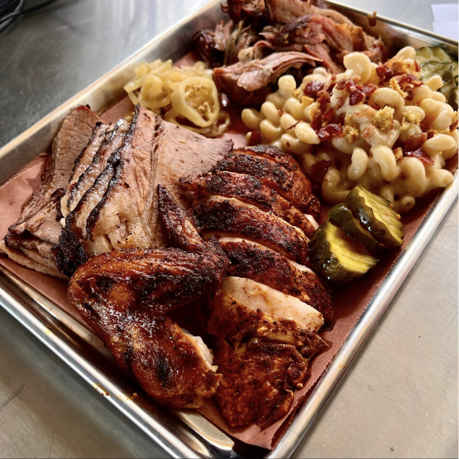 Close up of a BBQ platter with roasted chicken, Mac & Cheese, brisket, pickles on a metal tray. 