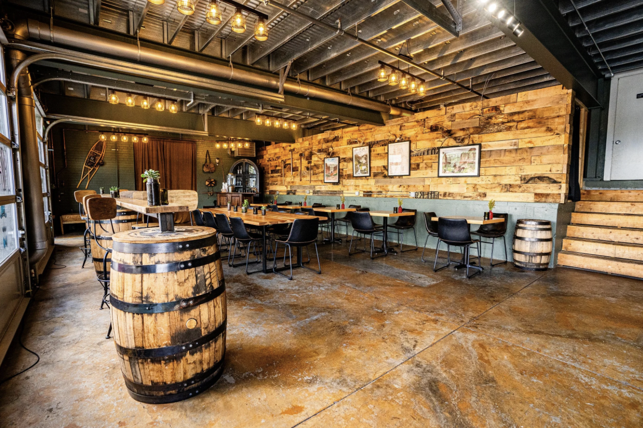 Interior of a rustic brewery or bar with wooden walls, black chairs around wooden tables, industrial lighting, and a small stage with chairs in the background.